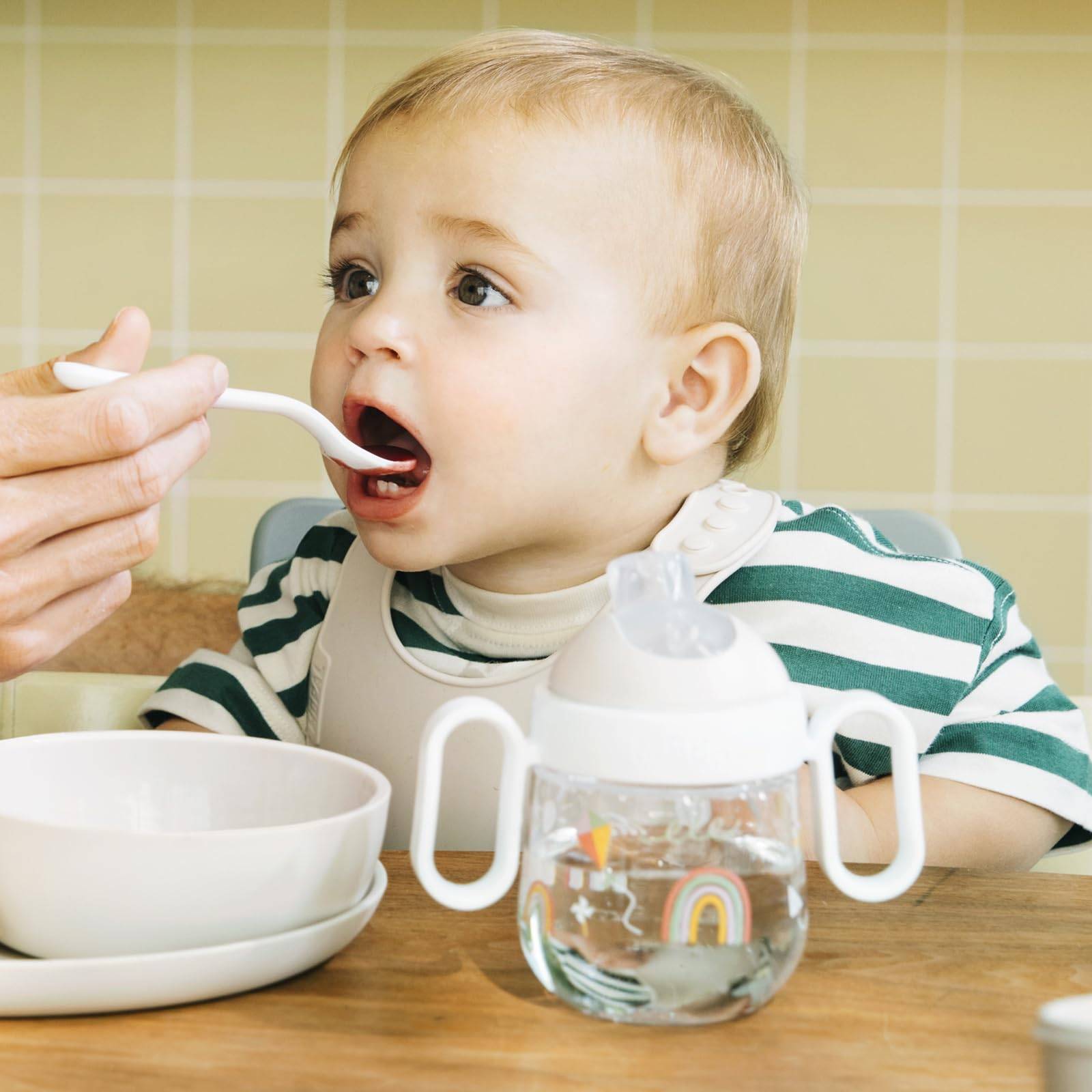 Ein Erwachsener füttert ein Baby mit einem Löffel an einem Tisch; eine Trinklernflasche und eine Schüssel befinden sich vor dem Kind.