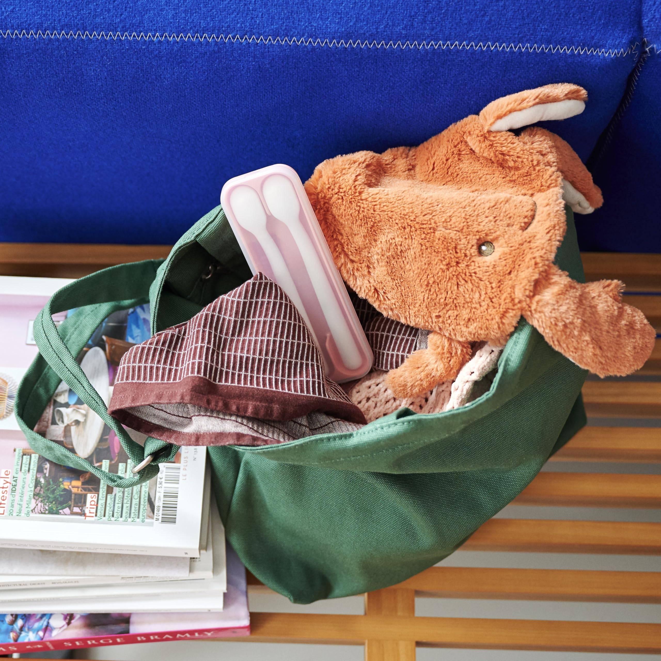 Grüne Einkaufstasche, gepackt mit Büchern, einem rosa Etui und einem orangefarbenen Plüschtier auf einem Tisch oder einer Bank.