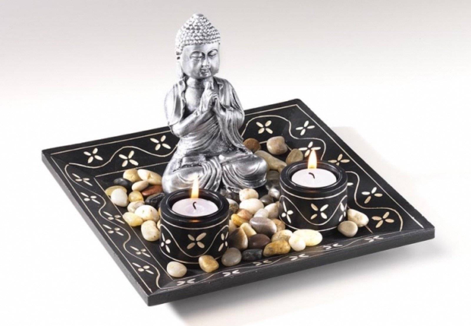 Dekorative Meditationsanzeige mit einer silbernen Buddha-Statue, zwei brennenden Kerzen, Kieselsteinen und einem gemusterten schwarzen Tablett.