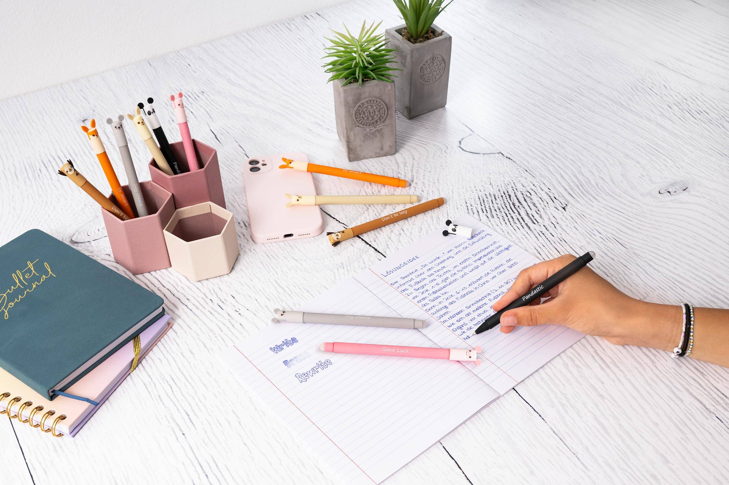 Handschriftliche Notizen auf liniertem Papier an einem ordentlichen weißen Schreibtisch mit Stiften, Bleistiften, Notizbüchern und kleinen Topfpflanzen.