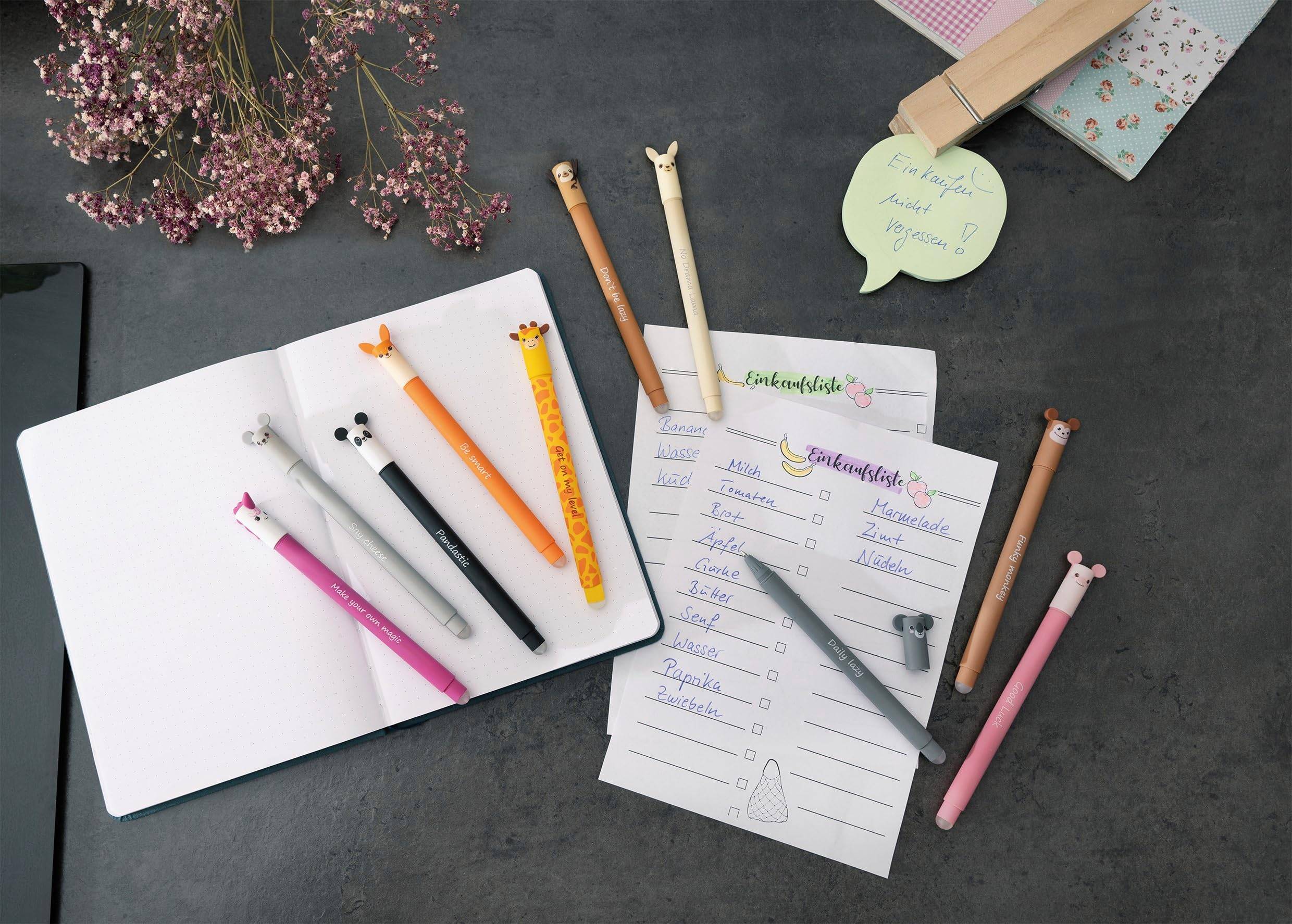 Notizbuch und To-Do-Liste verteilt mit bunten Stiften, Bleistiften und einem rosa Notizbuch auf einer dunklen Tischplatte, was auf Planung oder Tagebuchführung hindeutet.