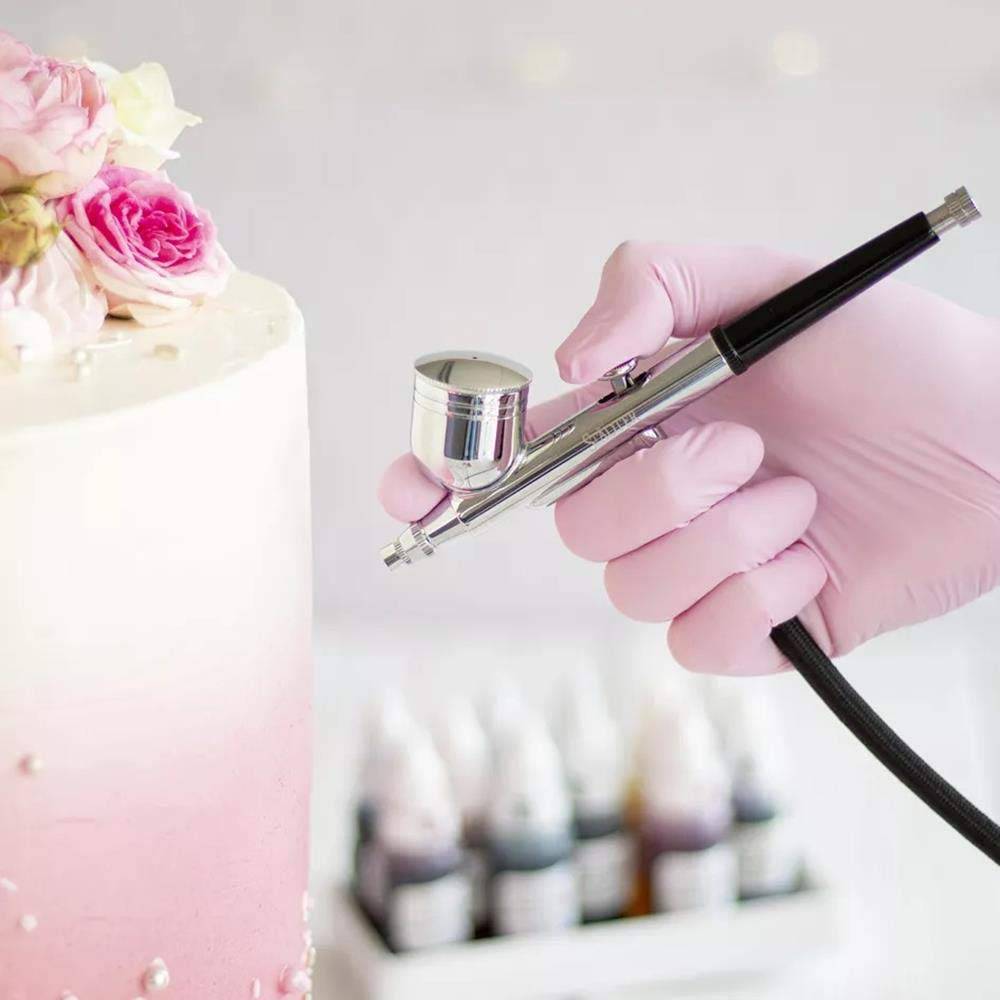 Behandschuhte Hand hält eine Airbrush auf einen rosa glasierten Kuchen mit Blumendekoration gerichtet, mit unscharfen Farbflaschen im Hintergrund.