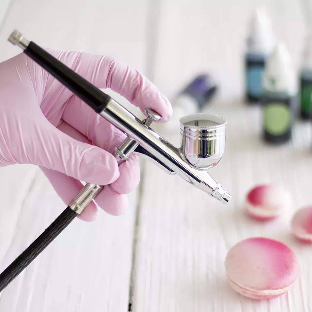 Eine behandschuhte Hand hält ein Airbrush-Gerät, das auf rosa Makronen auf einer weißen Oberfläche ausgerichtet ist, was auf Dekorations- oder Lebensmittelfärbungsarbeit hindeutet.