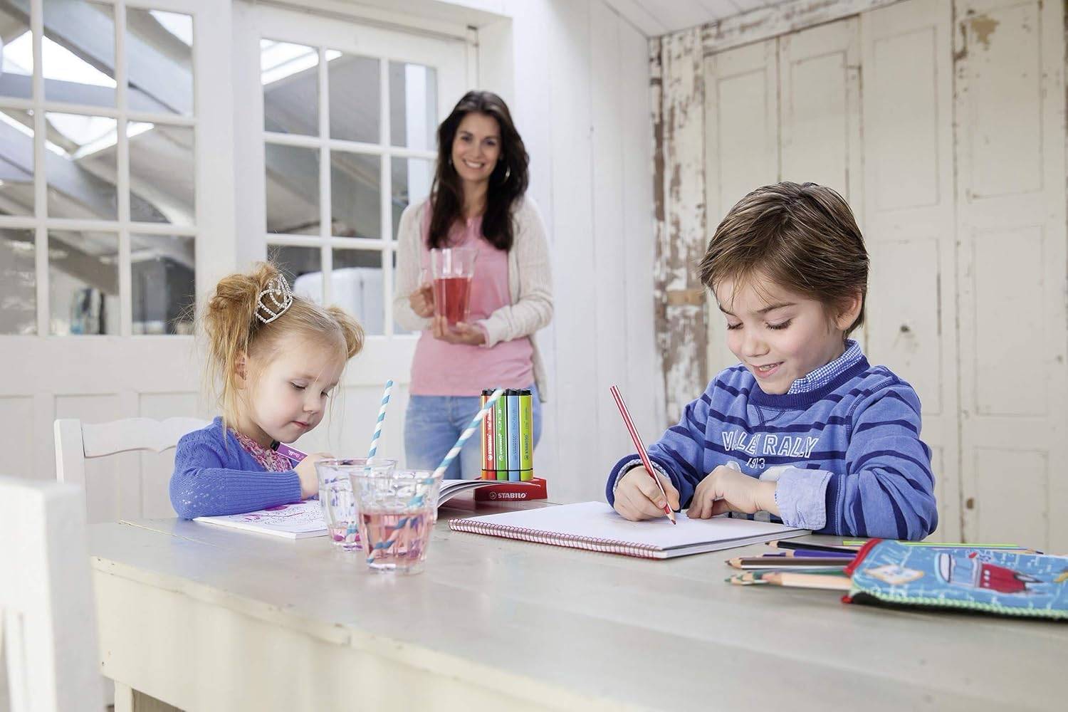 Zwei Kinder zeichnen an einem Tisch, während ein Erwachsener hinter ihnen steht und Getränke hält, in einer hellen Küche oder Esszimmer.