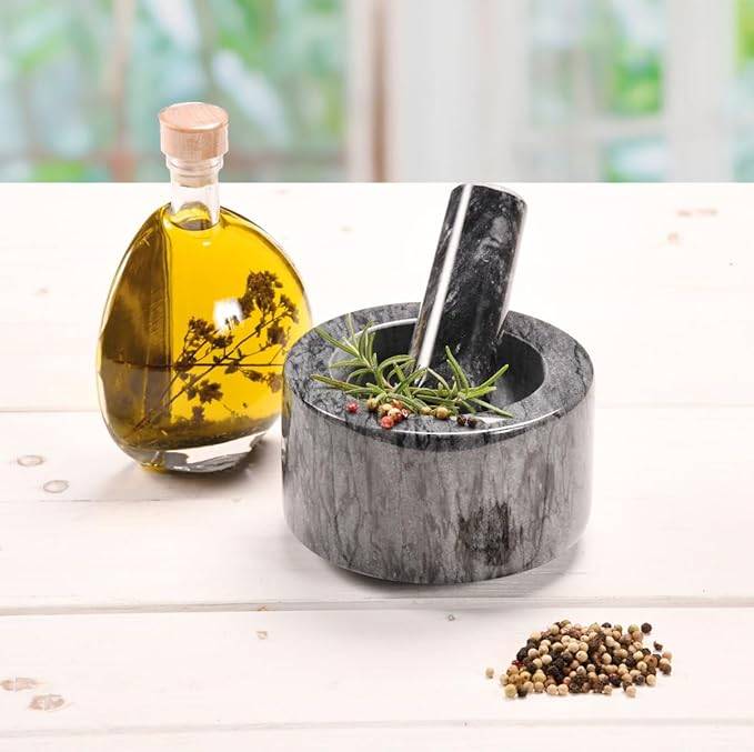 Marmormörser und Stößel mit Kräutern und Pfefferkörnern neben einer Flasche Olivenöl auf einem weißen Tisch.