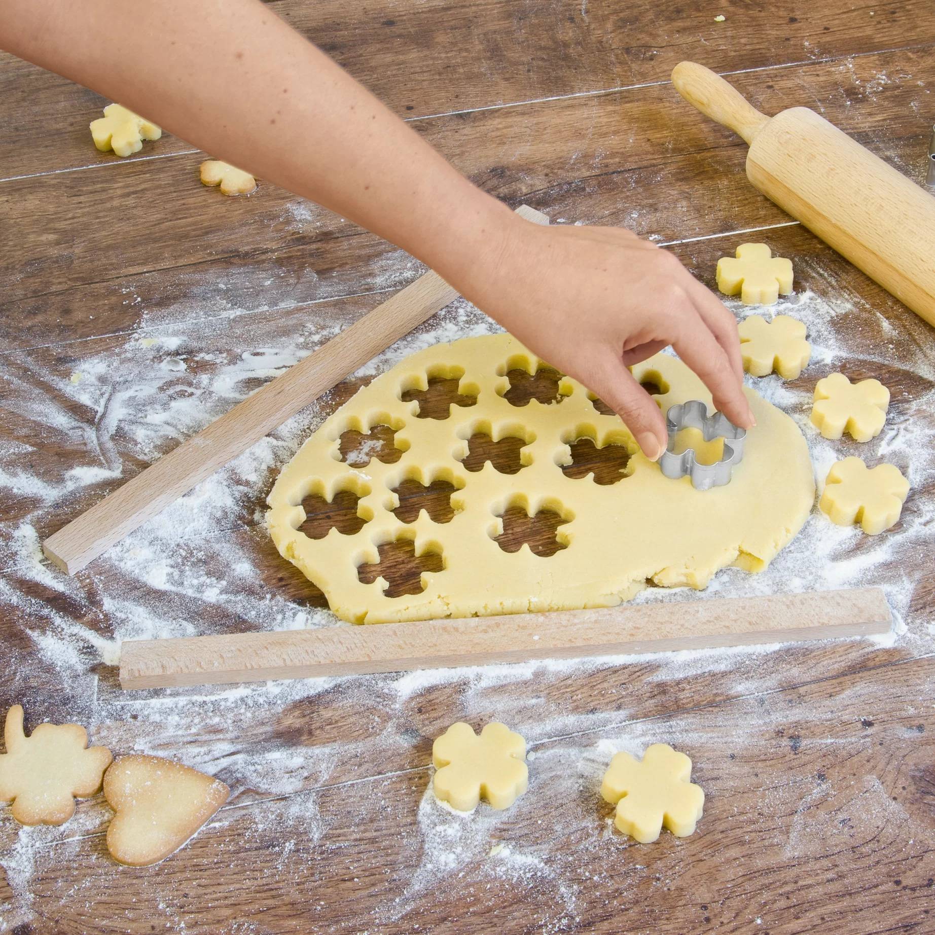 Kind verwendet einen blumenförmigen Plätzchenausstecher, um Teig auf einem bemehlten Holztisch auszustechen, mit Nudelholz und ausgestochenen Plätzchen in der Nähe.