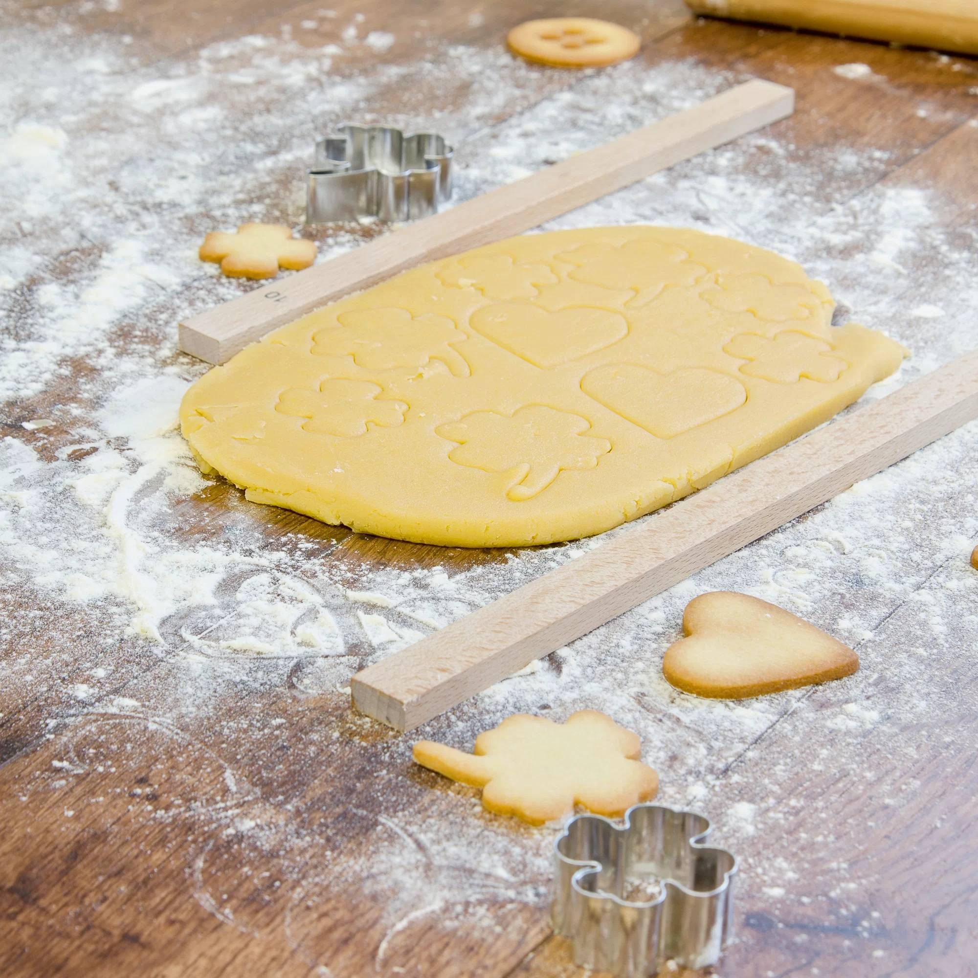 Ausgerollter Zucker-Plätzchenteig auf einer mehlbestäubten Holzoberfläche mit ausgestochenen Plätzchenformen, Metallausstechern und Holzstäbchen für gleichmäßige Dicke.