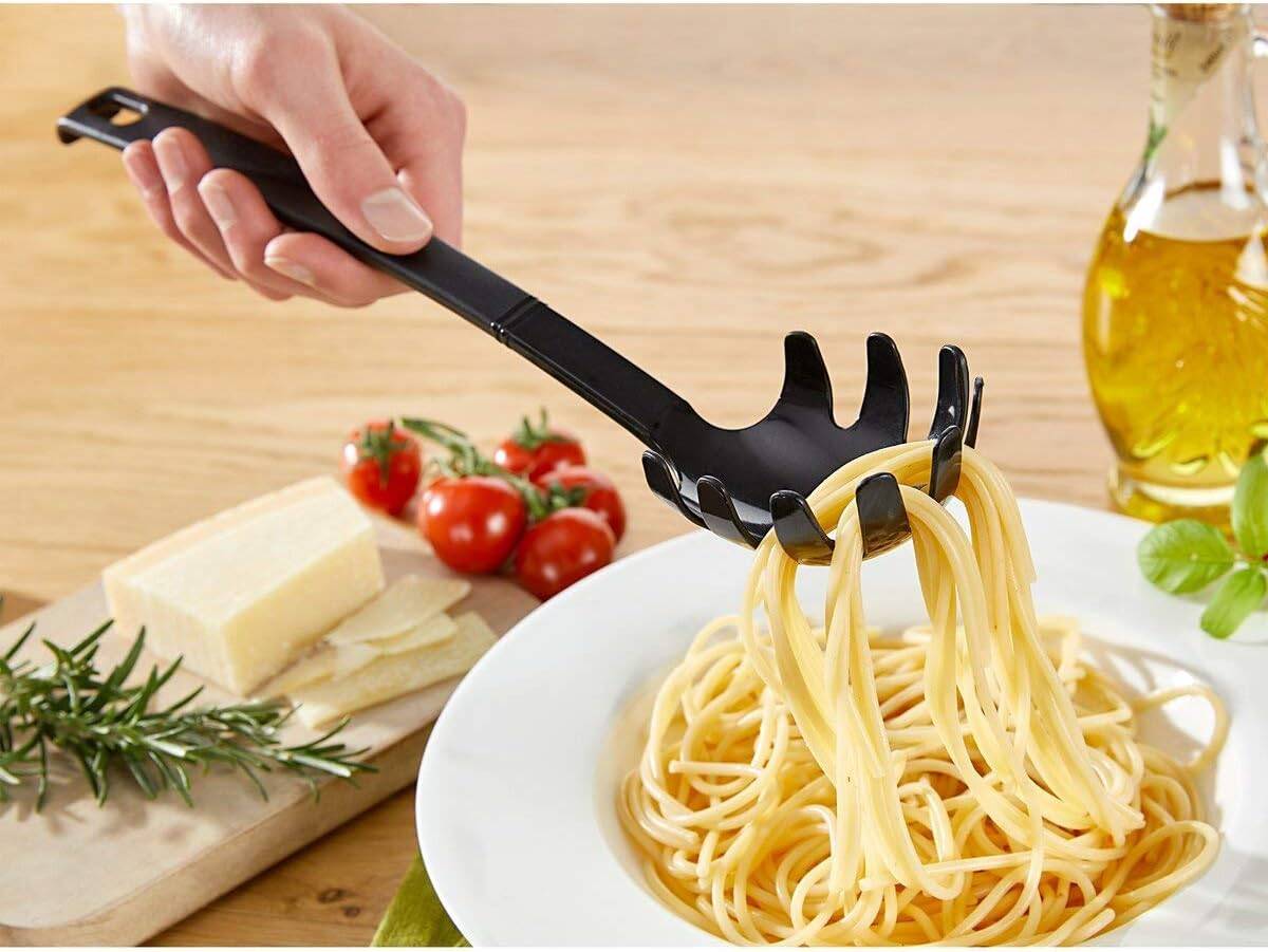 Hand benutzt einen schwarzen Pastaloeffel, um Spaghetti von einem weißen Teller zu heben, mit Tomaten, Käse, Kräutern und Olivenöl auf einem Holztisch.