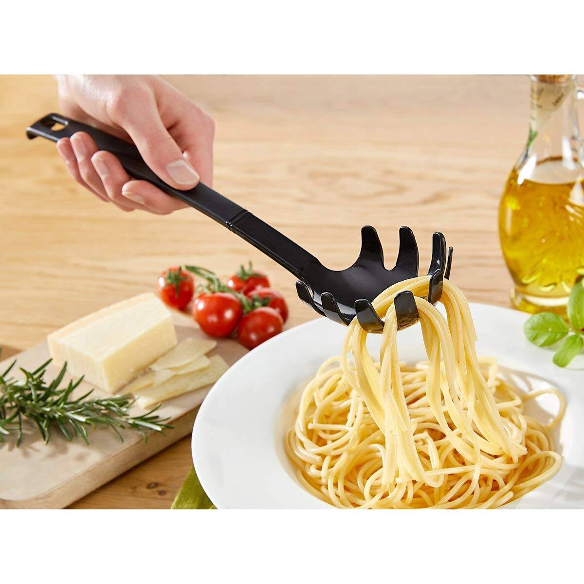 Eine Hand serviert Spaghetti mit einem schwarzen Pastaloeffel auf einem weißen Teller, mit Tomaten, Käse, Kräutern und Olivenöl in der Nähe auf einem Holztisch.
