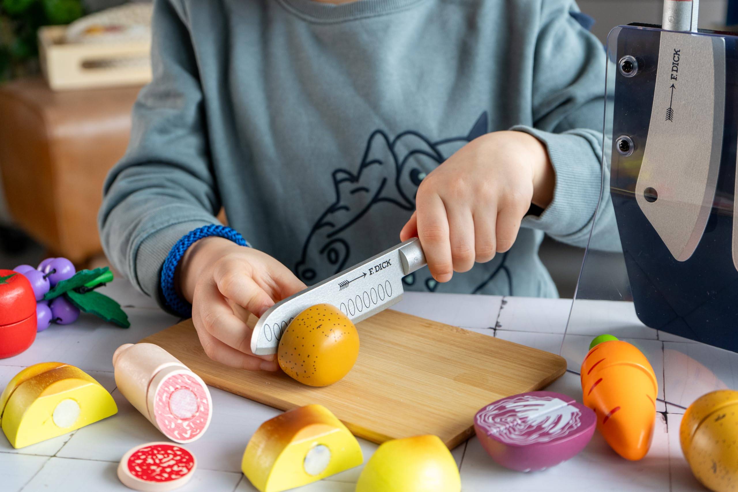 Kind, das mit einem Spielzeugmesser Spielzeugobst und -gemüse auf einem Schneidebrett während des Rollenspiels in einer küchen-ähnlichen Umgebung schneidet.