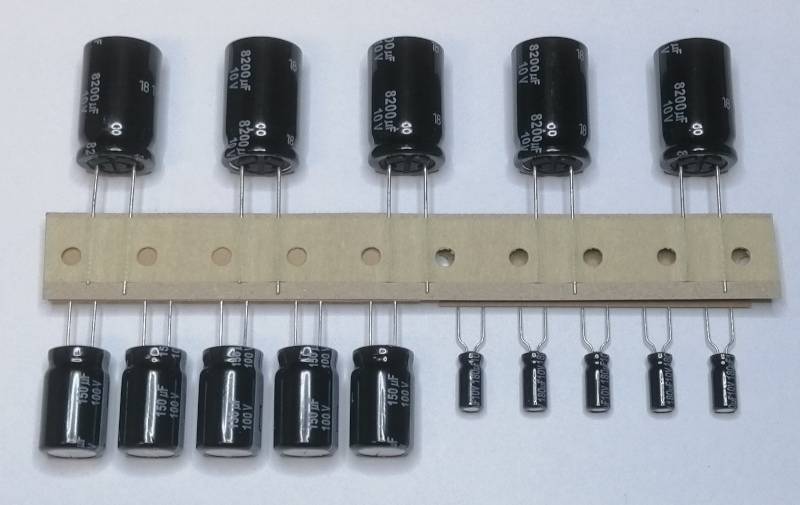 Verschiedene schwarze Elektrolytkondensatoren und kleinere Komponenten, arrangiert auf einem beigen Leiterplattenstreifen, gezeigt zum Größenvergleich und zur Montageplanung.