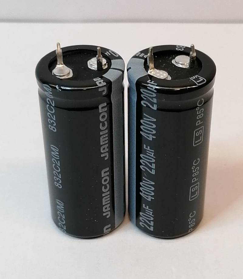 Zwei schwarze zylindrische Elektrolytkondensatoren mit Metallklemmen, aufrecht auf einer hellen Oberfläche stehend.