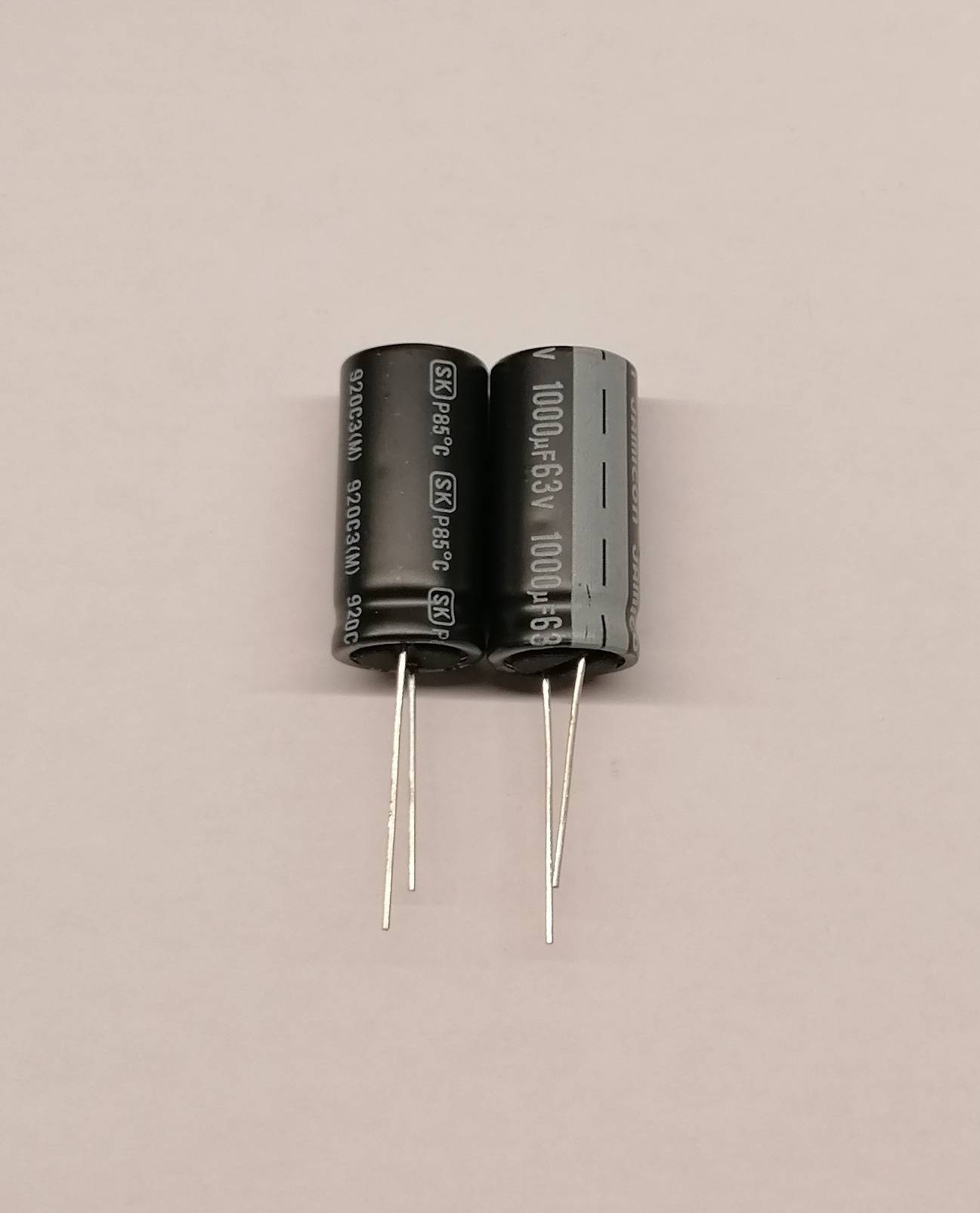 Zwei zylindrische Elektrolytkondensatoren nebeneinander auf einem hellenen Untergrund stehend, mit Metalleanschlüssen, die nach unten führen.