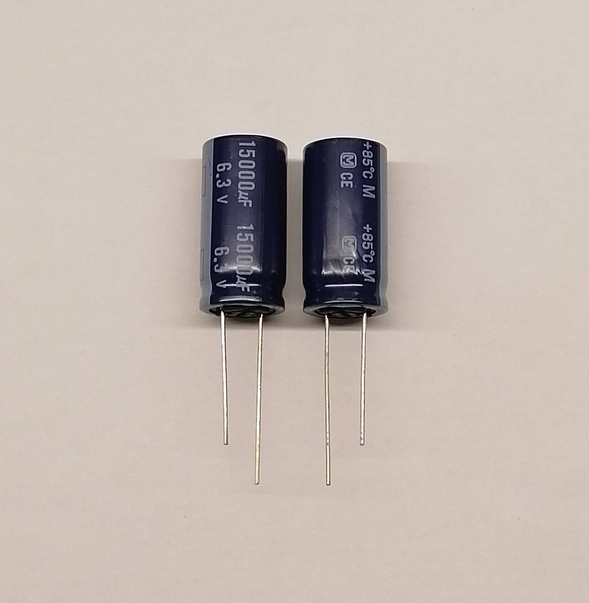 Zwei schwarze Elektrolytkondensatoren mit der Beschriftung 15000µF 6,3V, aufrecht stehend mit Metalldrähten auf einer ebenen Fläche.