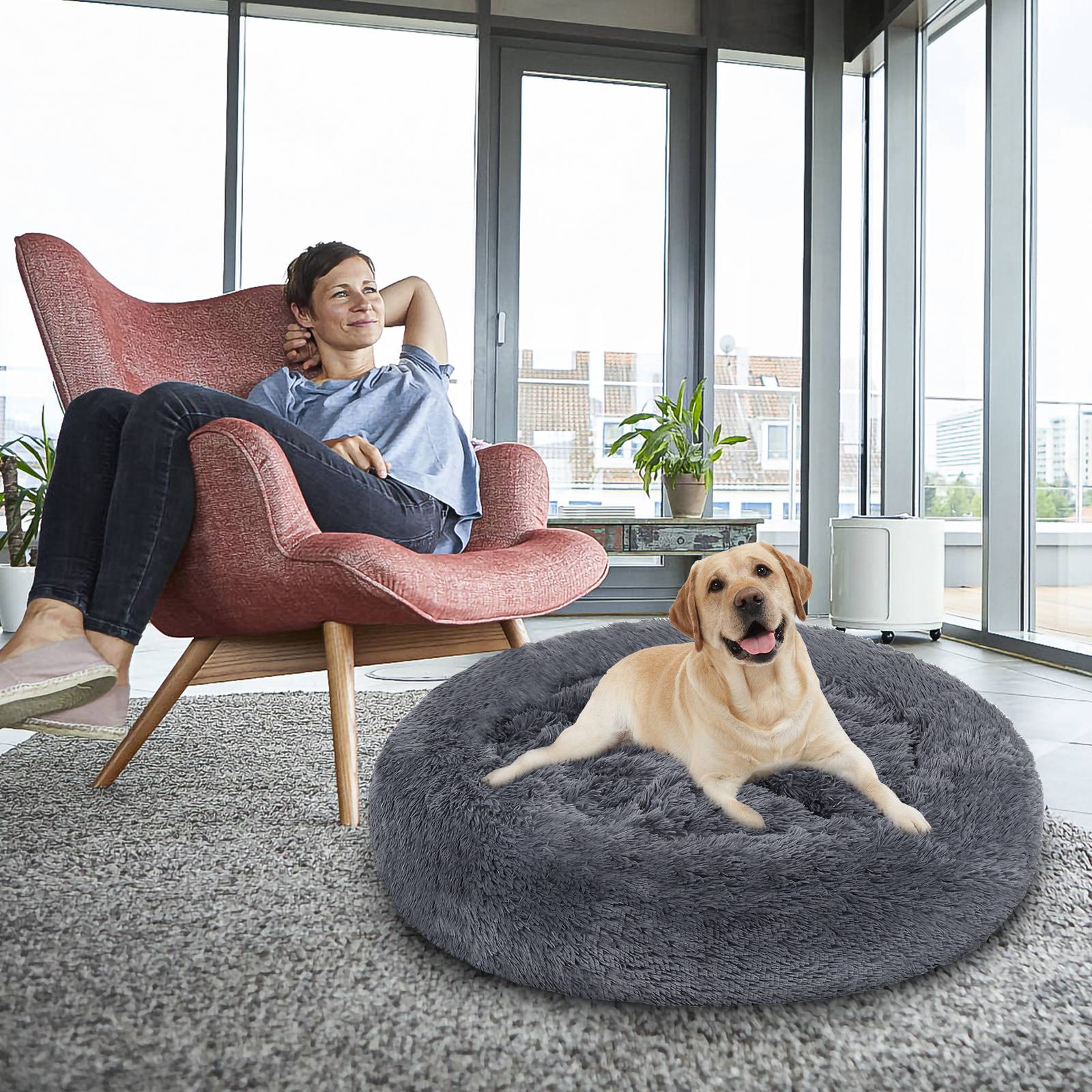 Eine Frau entspannt sich in einem rosa Sessel neben einem gelben Hund, der auf einem grauen Haustierbett liegt, in einem hellen modernen Wohnzimmer.