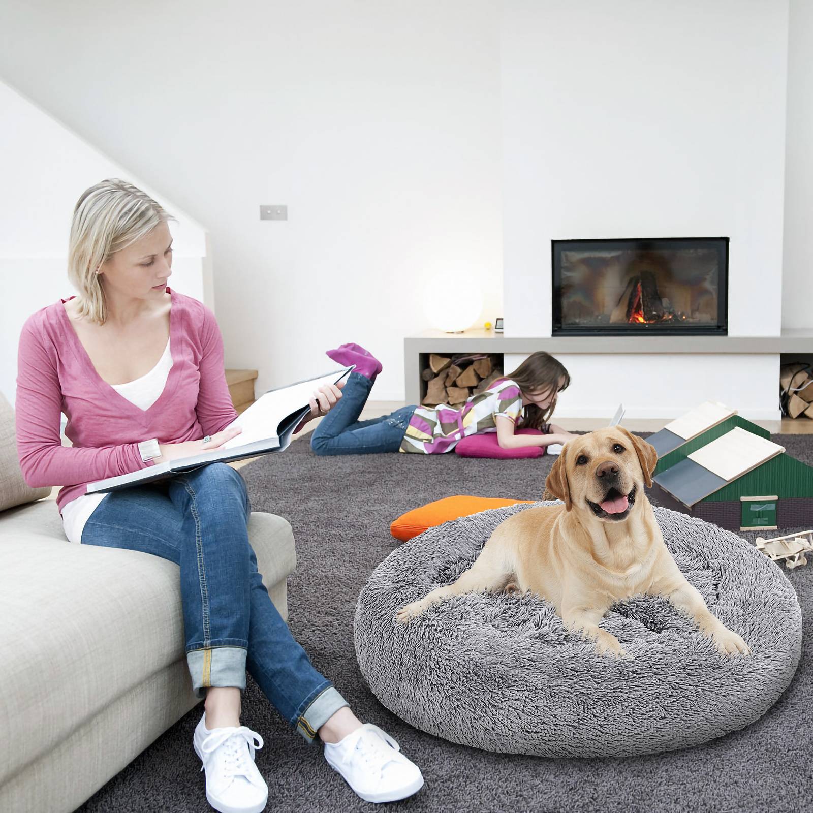 Eine Frau liest auf einem Sofa, während ein Kind auf dem Boden liegt und ein Hund auf einem Sitzsack in einem hellen Wohnzimmer mit einem Kamin ruht.