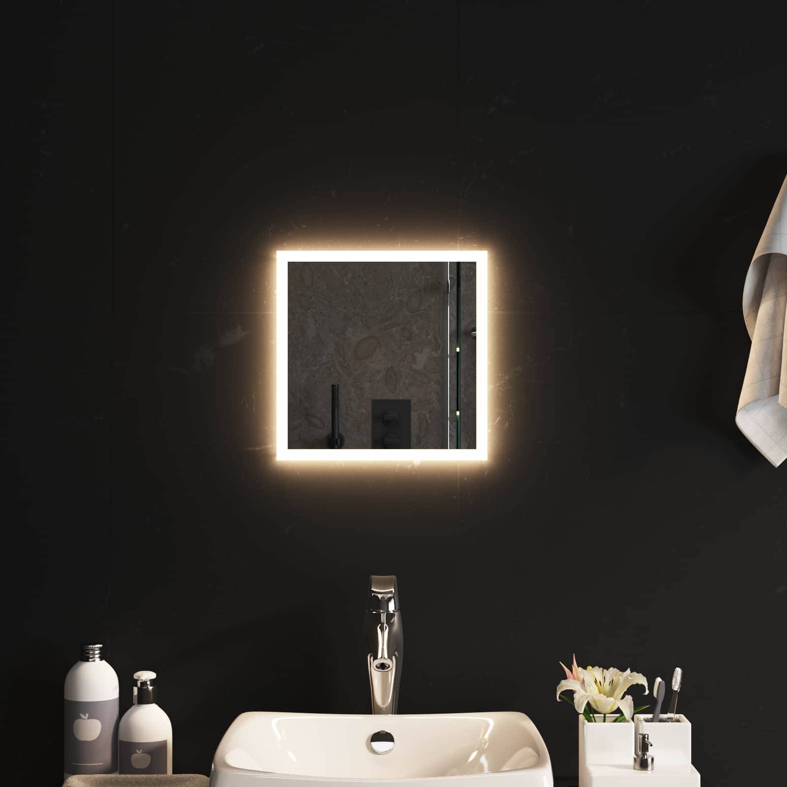 Modernes Badezimmermöbel mit beleuchtetem quadratischem Spiegel über einem weißen Waschbecken, flankiert von Seifenflaschen und einer kleinen Blumenarrangement, vor einer dunklen Wand.