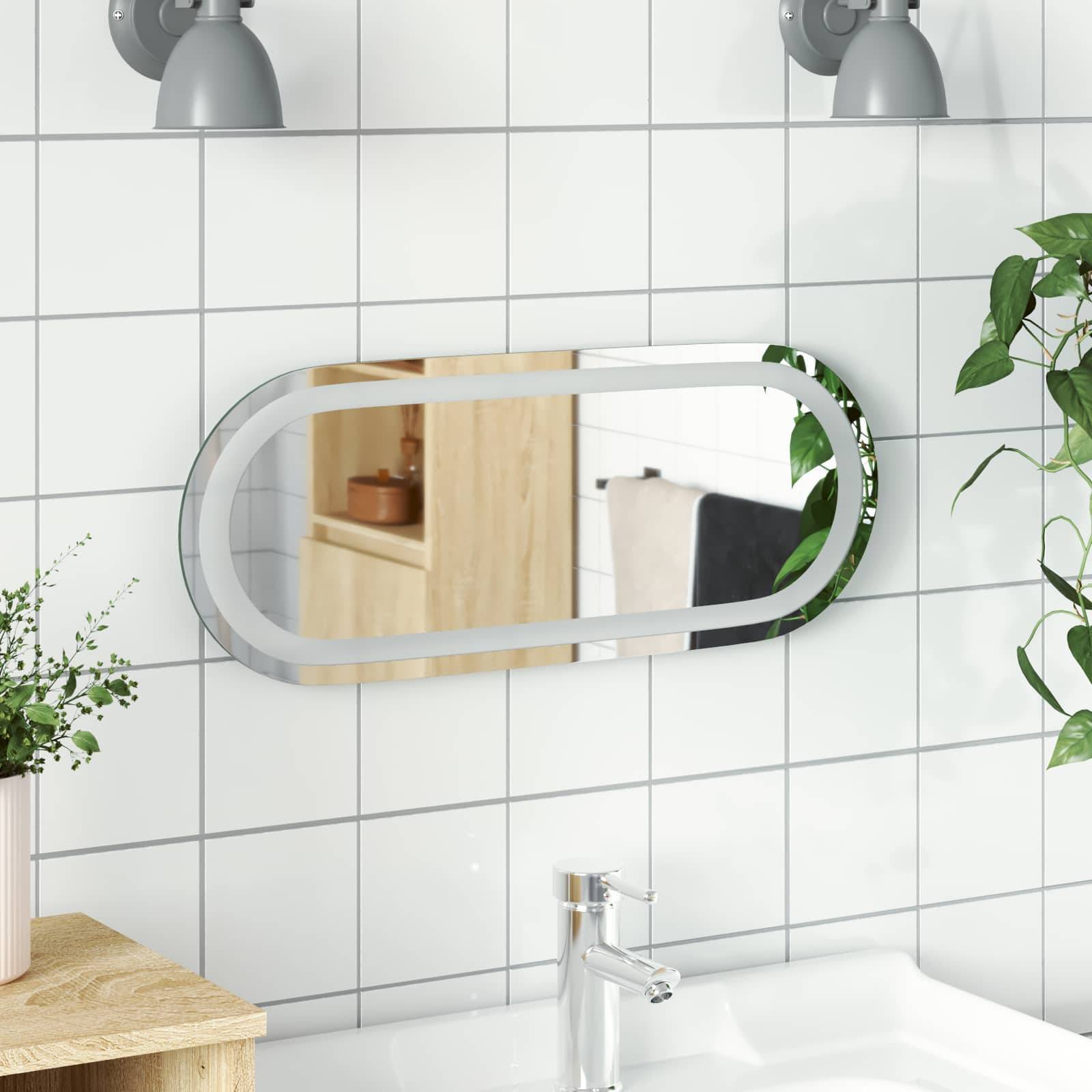 Modernes Badezimmer mit ovalem Wandspiegel über weißem gefliesten Waschbecken und Chromarmatur, mit Pflanzen und einem Handtuch, die sich im Spiegel spiegeln.