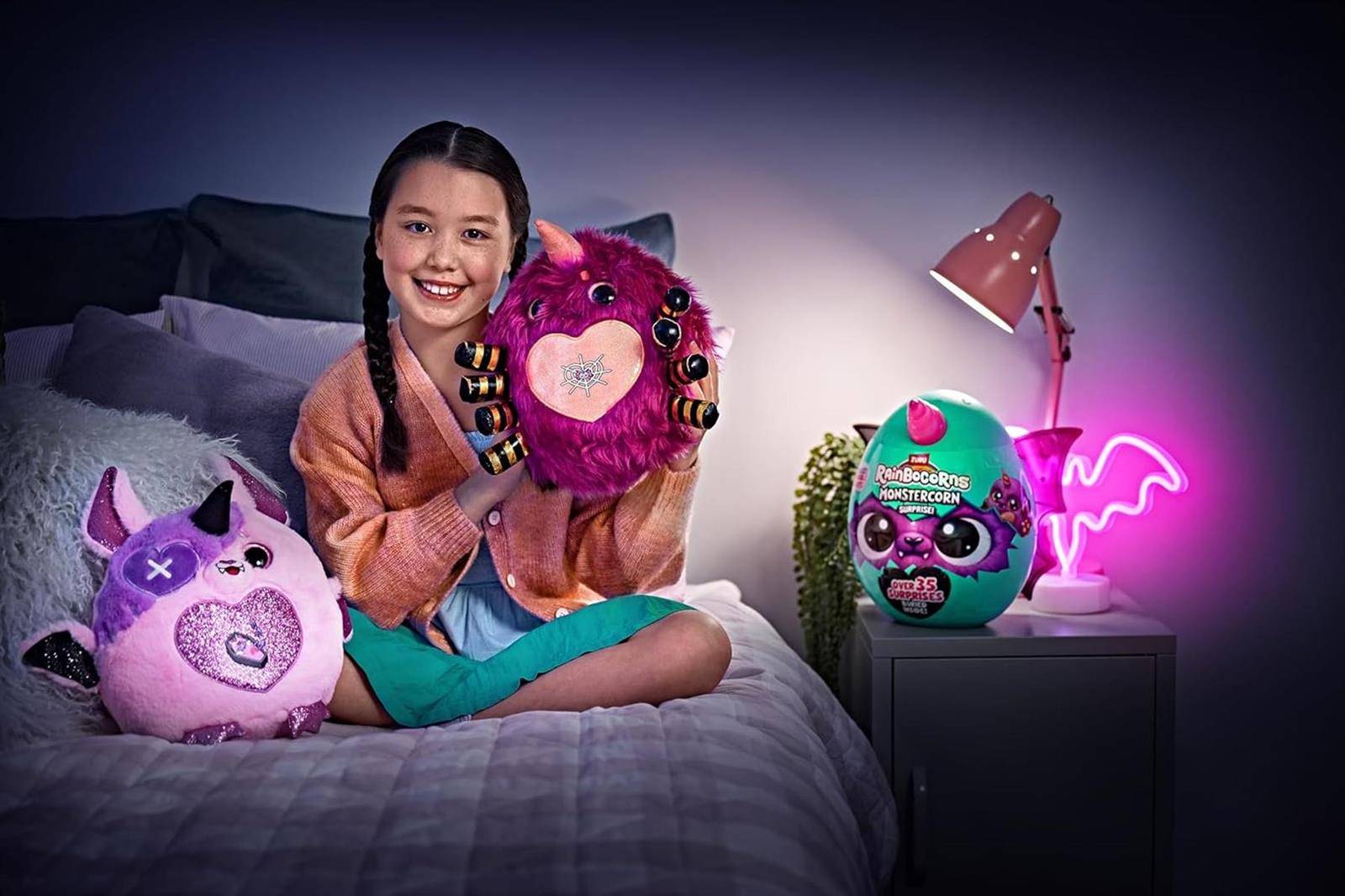 Mädchen, das auf einem Bett sitzt und ein rosa flauschiges Spielzeug hält, mit anderen Plüschtieren und einer Produktschachtel auf einem Nachttisch neben einer Lampe und einem Neon-Fledermaus-Schild.