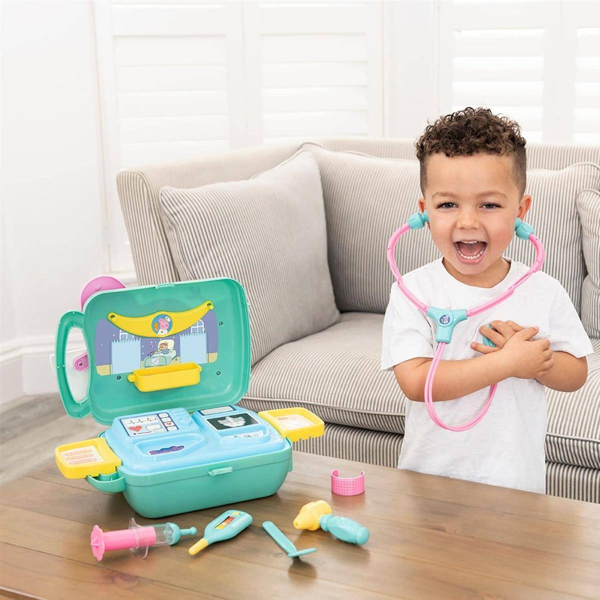 Lachendes Kind in einem Spielzeug-Arztoutfit hält ein Stethoskop neben einem offenen medizinischen Spielset auf einem Tisch im Wohnzimmer.