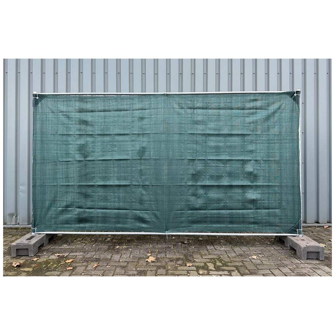 Grünes Mesh-Sichtschutzgewebe, das zwischen Metallpfosten vor einer gewellten Wand gespannt ist, mit Betonblöcken an der Basis.