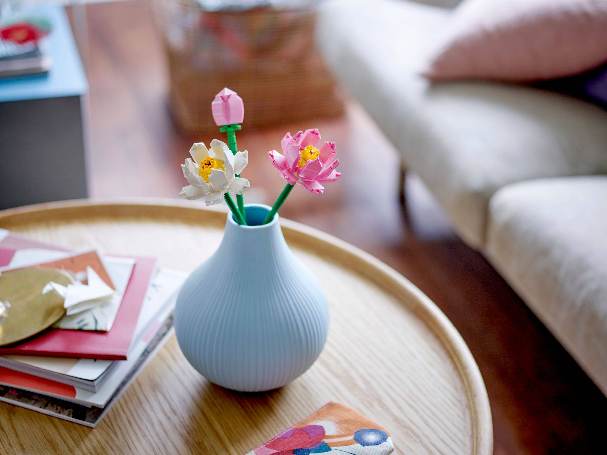 Blaue Vase mit drei Papierblumen auf einem runden Holz-Couchtisch neben gestapelten Zeitschriften und einem Sofa in einem Wohnzimmer.