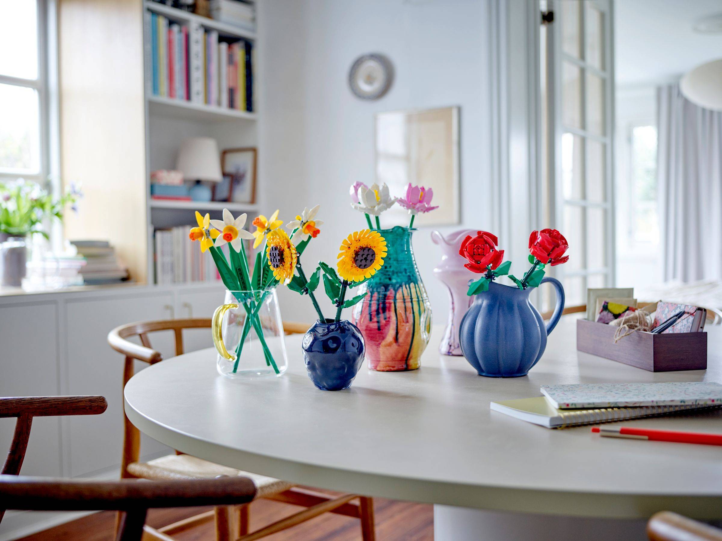 Farbenfrohe Papierblumenarrangements in Vasen auf einem weißen Tisch, mit einem Notizbuch, Bleistiften und einer kleinen Box in einem hellen Wohnungsinneren.
