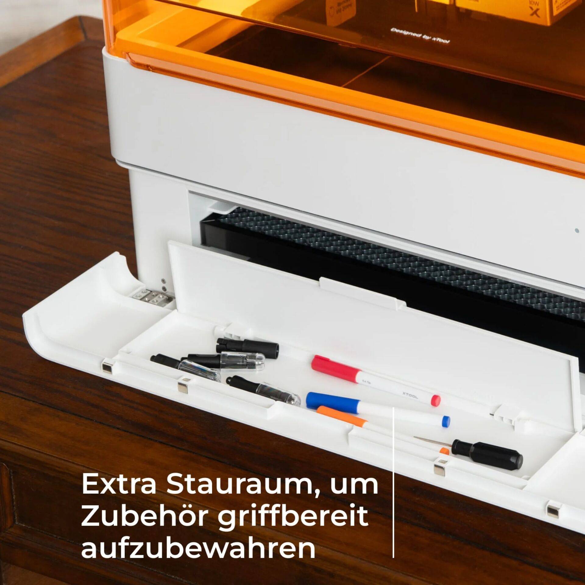 Schublade an einem druckerartigen Gerät mit Stiften und Markern öffnen; deutscher Text besagt „Extra Staurraum, um Zubehör griffbereit aufzubewahren