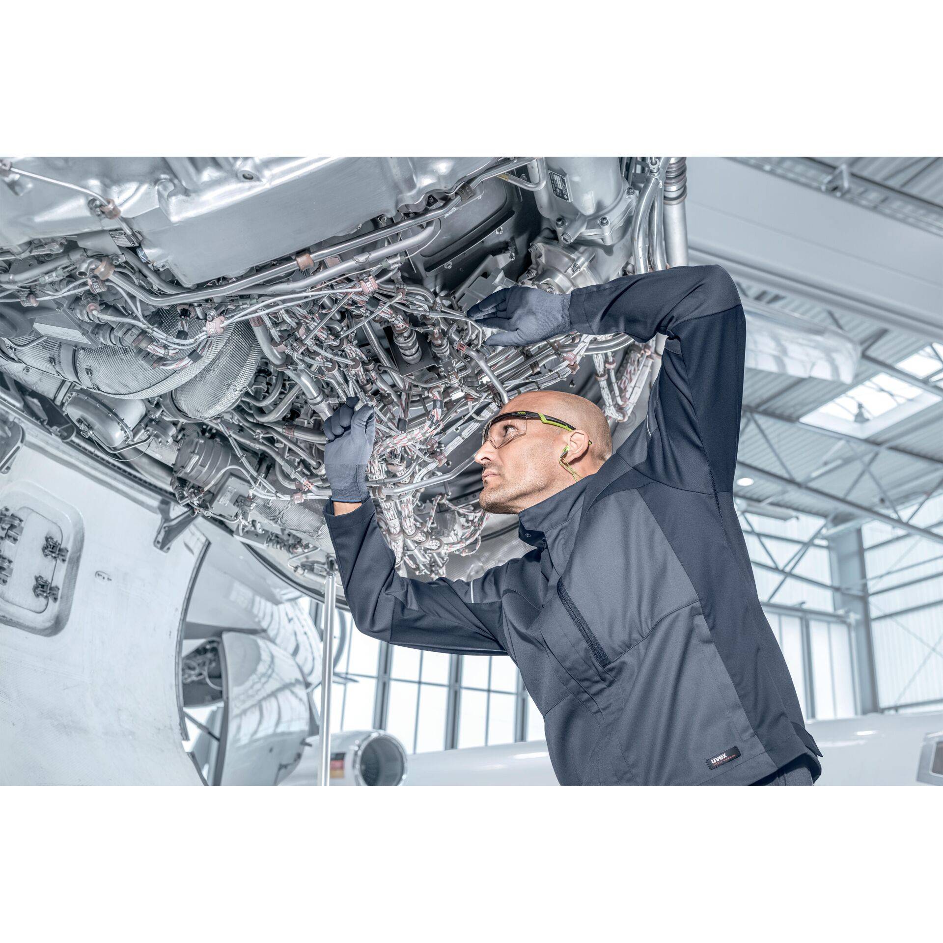 Techniker inspiziert komplexe Flugzeugverkabelung und Komponenten unter einem Flugzeug in einer hellen Wartungshalle.