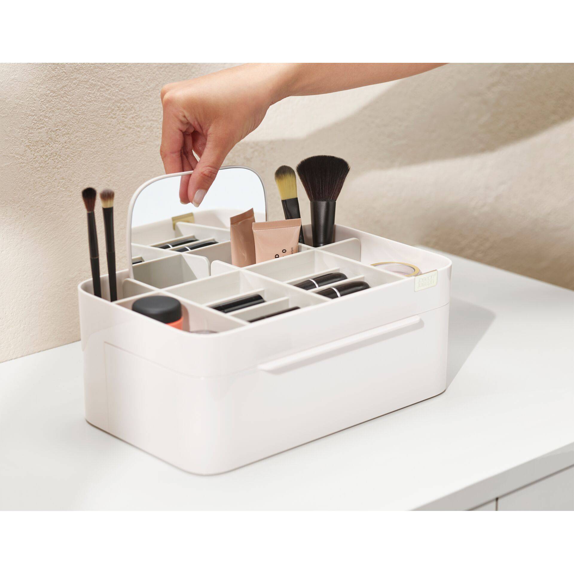 Hand, die einen Spiegel in einen weißen Make-up-Organizer legt, der mit Pinseln und Kosmetikprodukten auf einem Tisch gefüllt ist.
