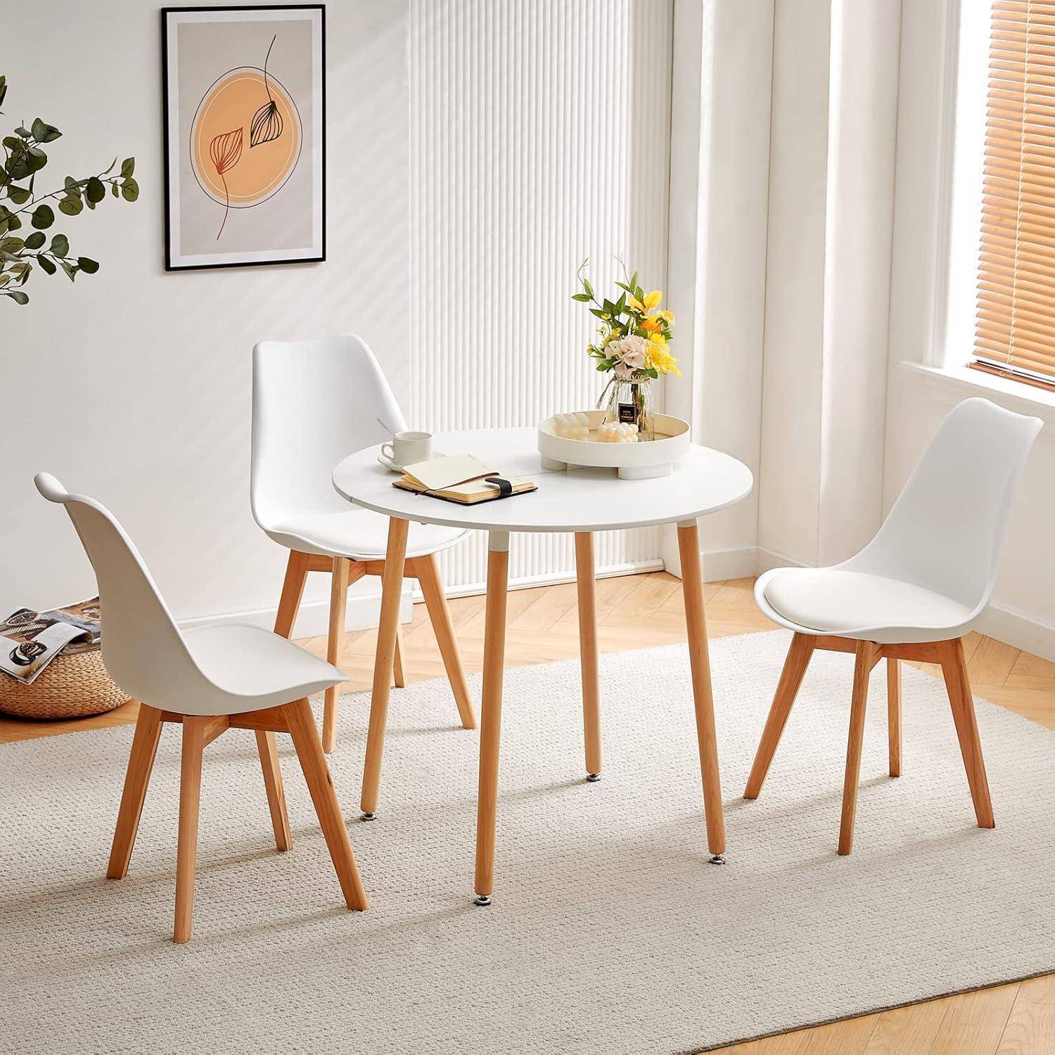 Moderner Essbereich mit einem runden weißen Tisch, drei weißen Stühlen und einer kleinen Blumenvase als Tischdekoration in einem hellen Raum mit Teppich und Wandkunst.