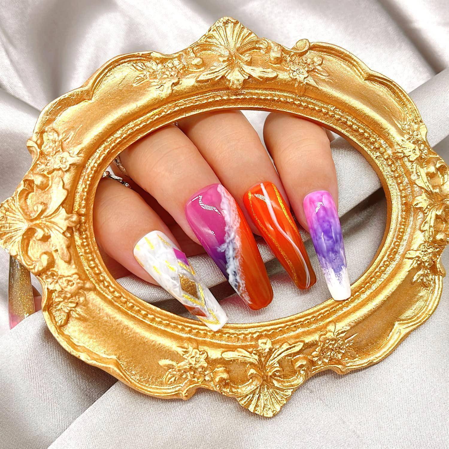 Hand mit langen, spitzen mehrfarbigen Marmor-Nageldesigns, gerahmt von einem verzierten goldenen ovalen Spiegel auf weißem Satinstoff.