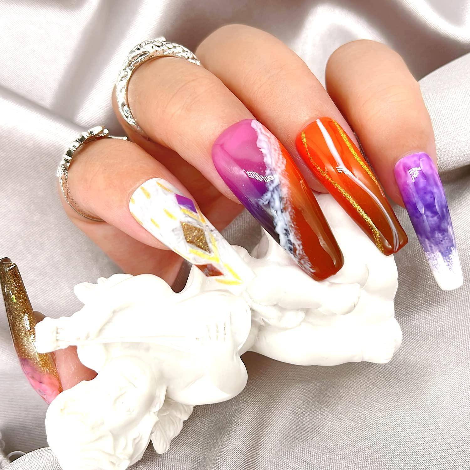 Hand zeigt lange, bunte gemusterte Acrylnägel mit metallischen und marmorierten Designs, gezeigt vor einem hellen Satinhintergrund.