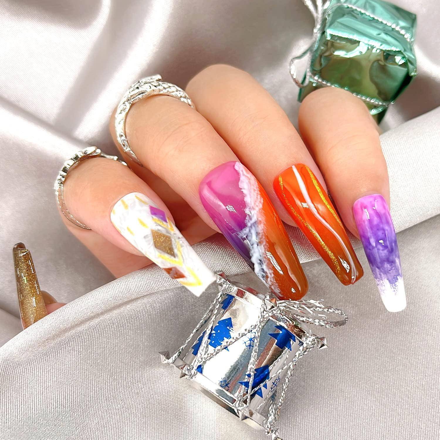 Hand mit langen, farbenfrohen Acrylnägeln in Rosa, Orange, Lila und Weiß mit abstrakten Designs, über Satinstoff mit metallischen Geschenk-ähnlichen Akzenten posiert.