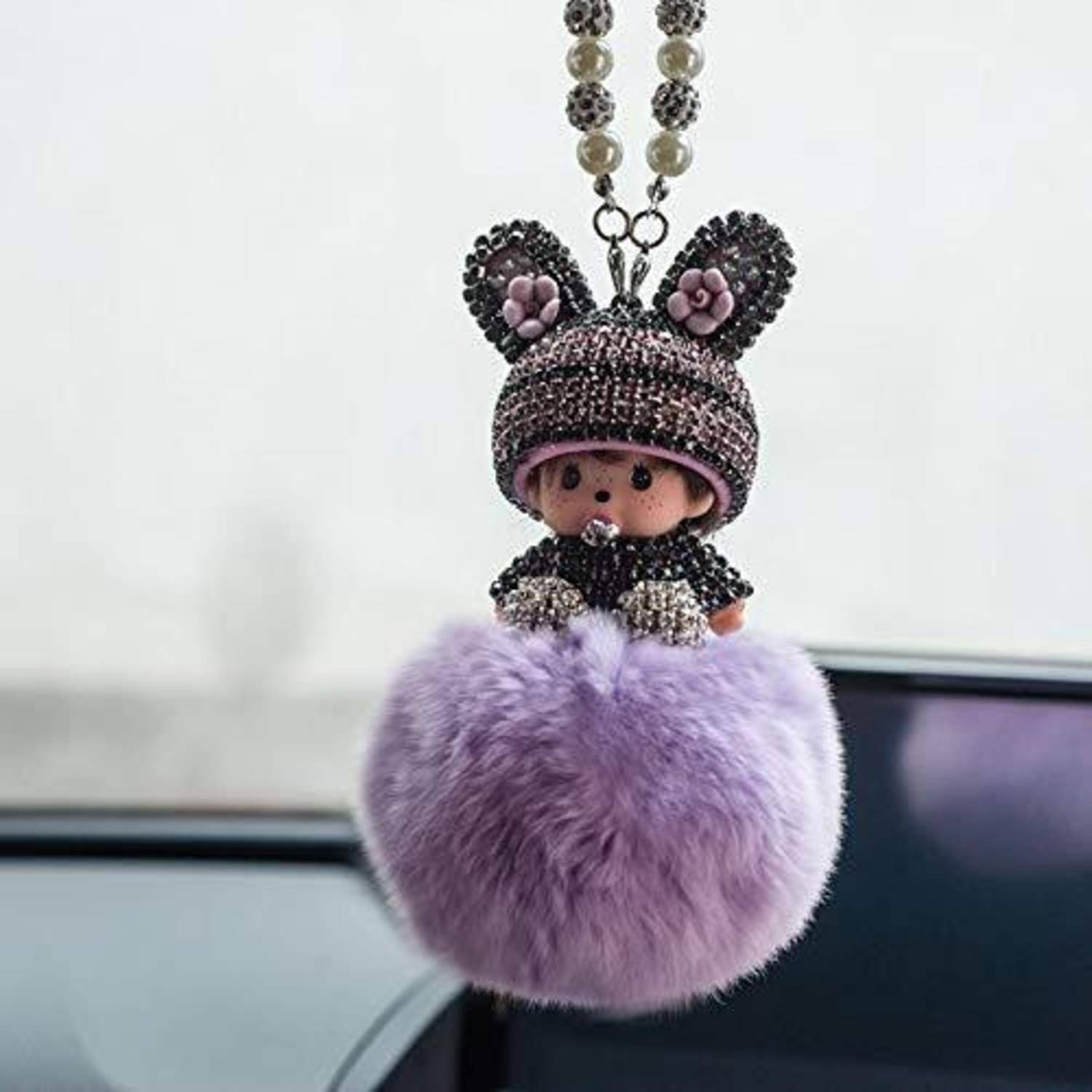 Eine Schlüsselanhänger-Puppe aus Perlen mit Mausohren und einem flauschigen Bommel-Körper in Lavendelfarbe, das in einem Autoinnenraum hängt.