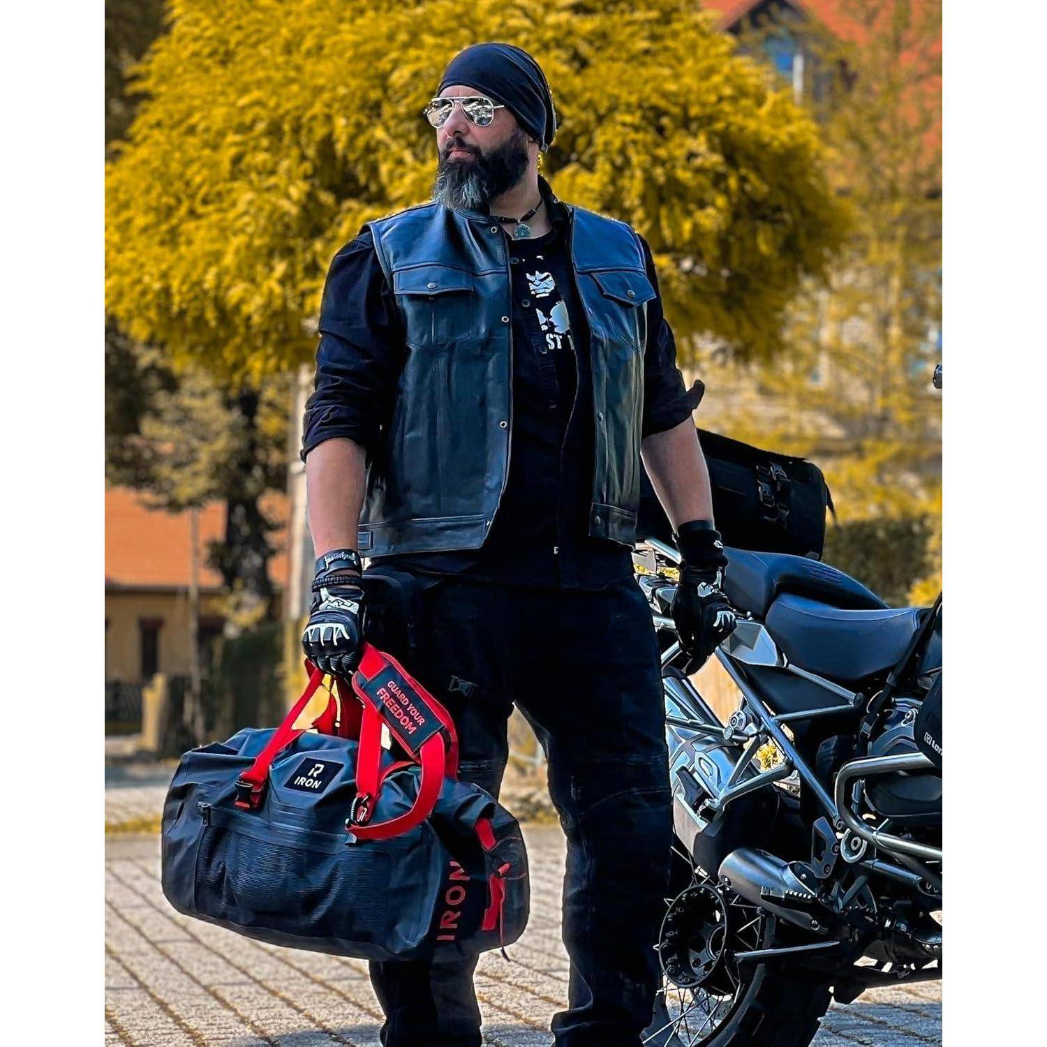 Ein bärtiger Mann in Motorradbekleidung trägt eine Reisetasche und steht neben einem Motorrad auf einer gepflasterten Straße im Freien mit Bäumen im Hintergrund.
