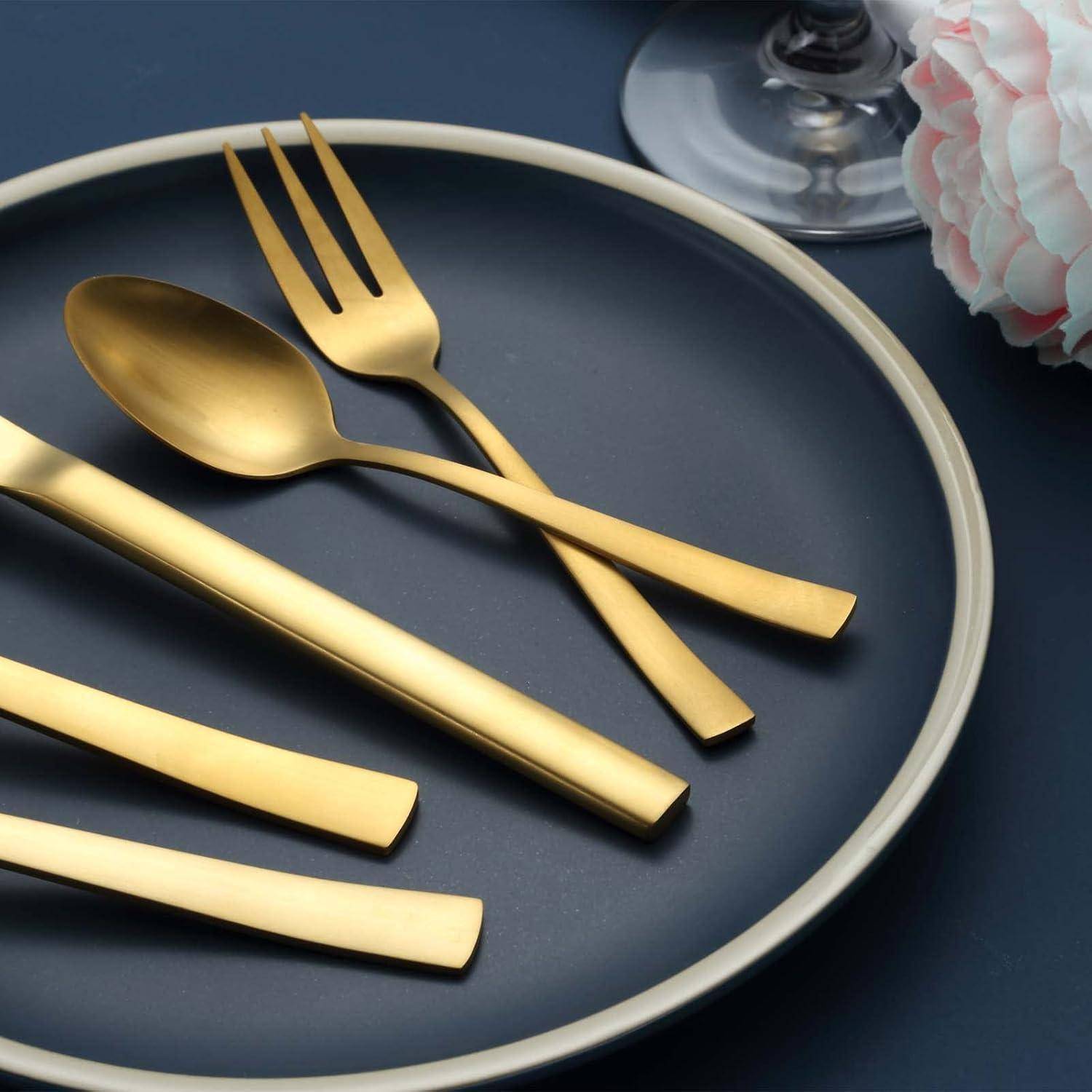 Goldfarben Löffel, Gabel und Messer auf einem dunklen Teller angeordnet, mit einem Glas und einer Blume in der Nähe.
