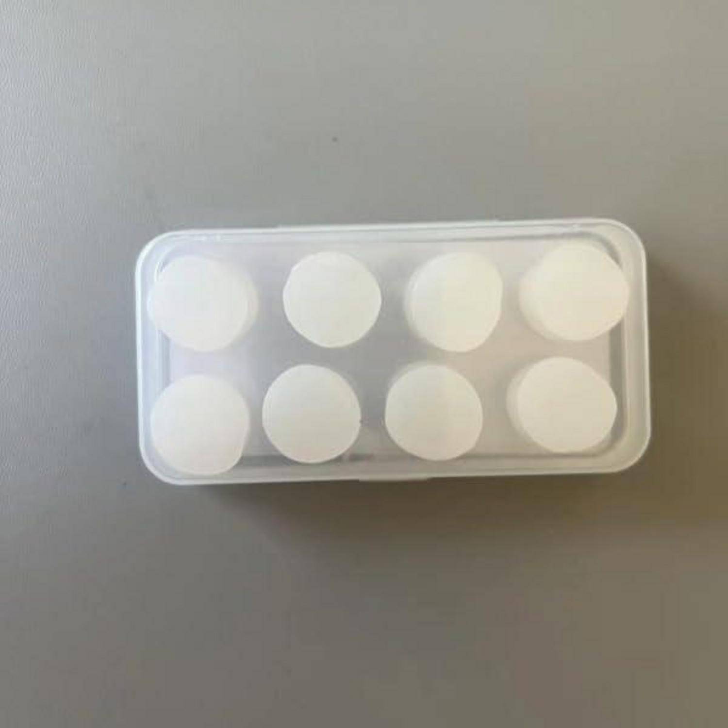 Kleine transparente Kunststoffschachtel mit acht weißen runden Tabletten auf einer grauen Oberfläche.