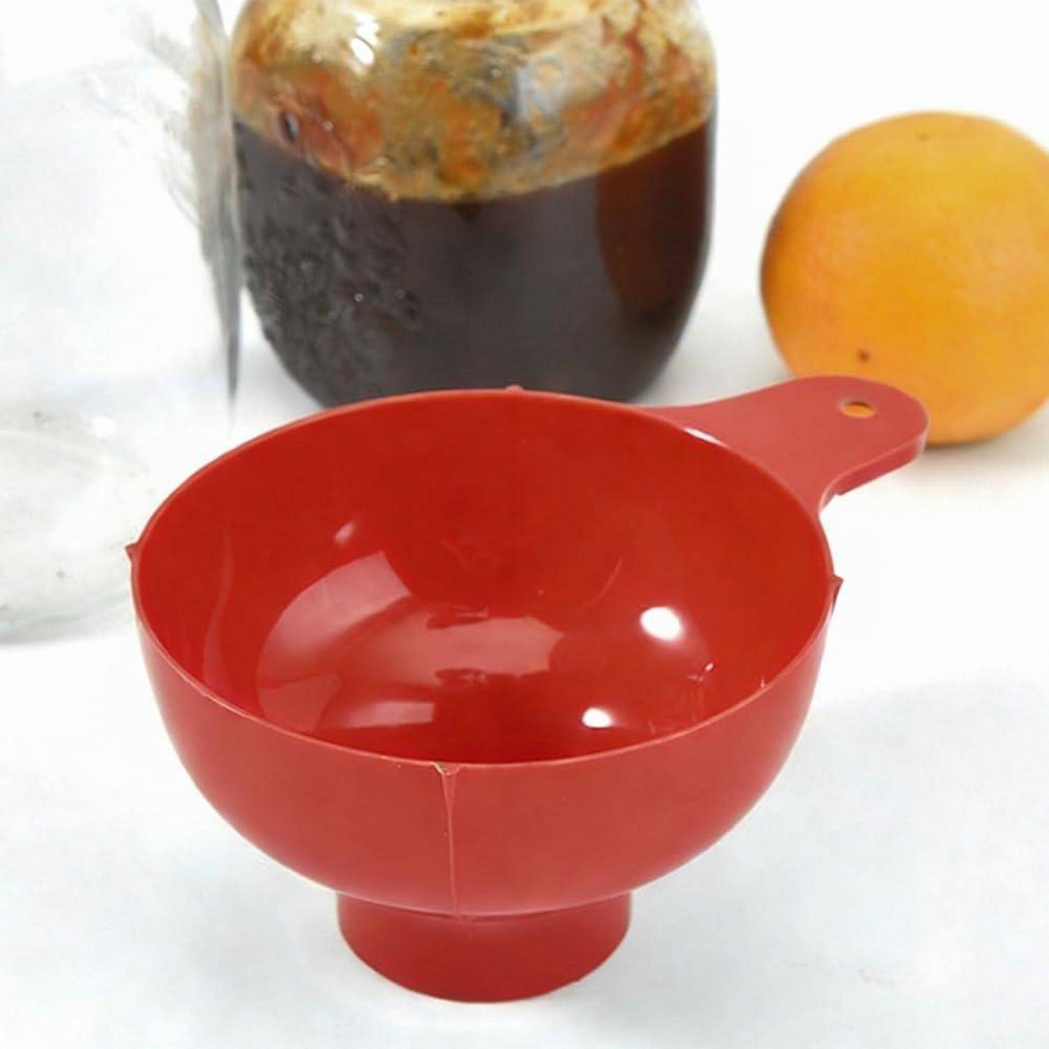 Rotes Kunststoff-Messfunnelbecher mit Griff auf einer weißen Oberfläche, mit einem Glas und einer Orange im Hintergrund.
