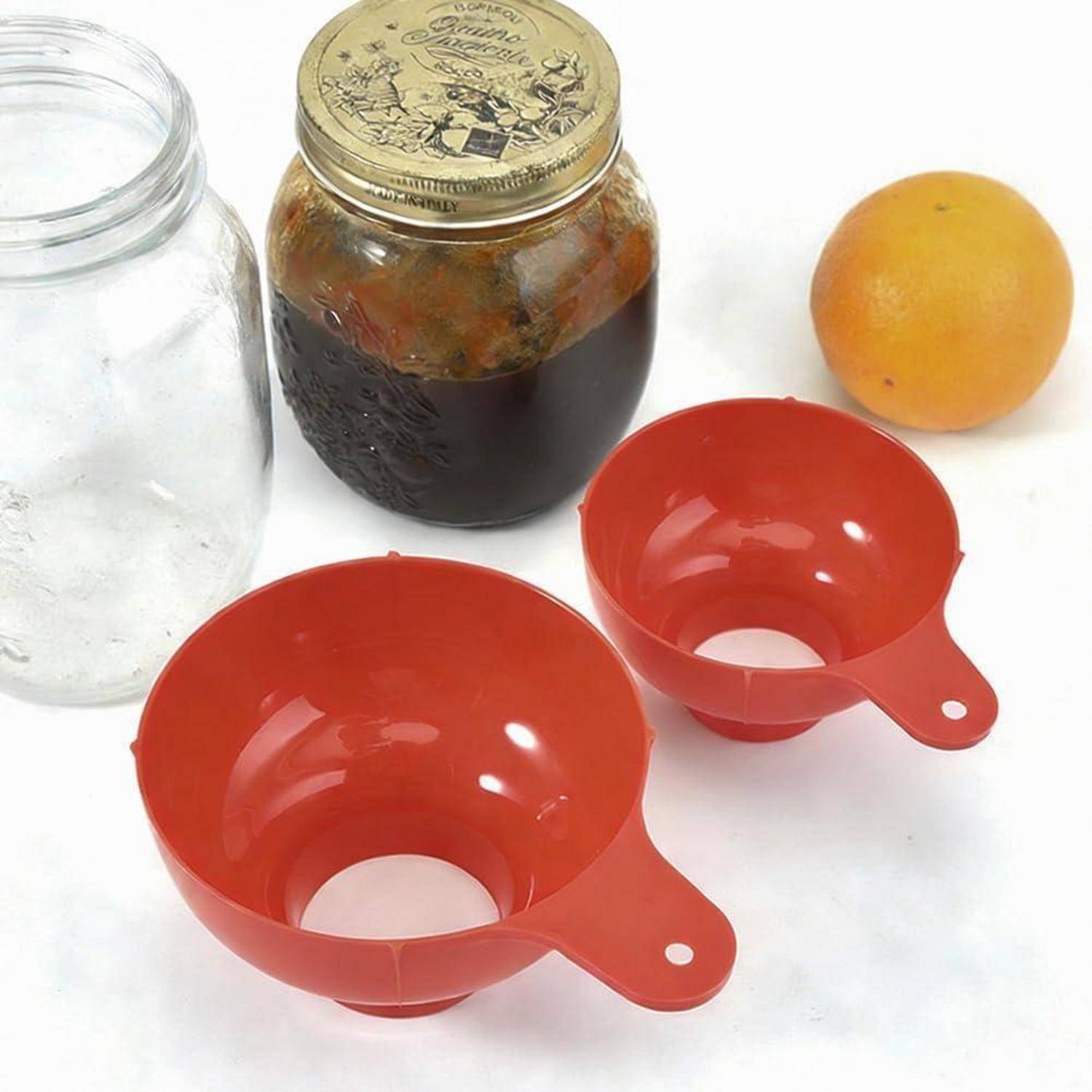 Zwei rote Kunststofftrichter, ein Glasgefäß mit Metalldeckel, ein leeres Glas und eine Orange auf einer weißen Oberfläche.