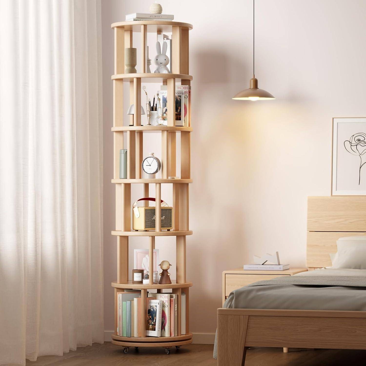 Hohes Holz-Drehbücherregal in einem Schlafzimmer mit Büchern und Dekorationen neben einem Bett und Fenstervorhängen.