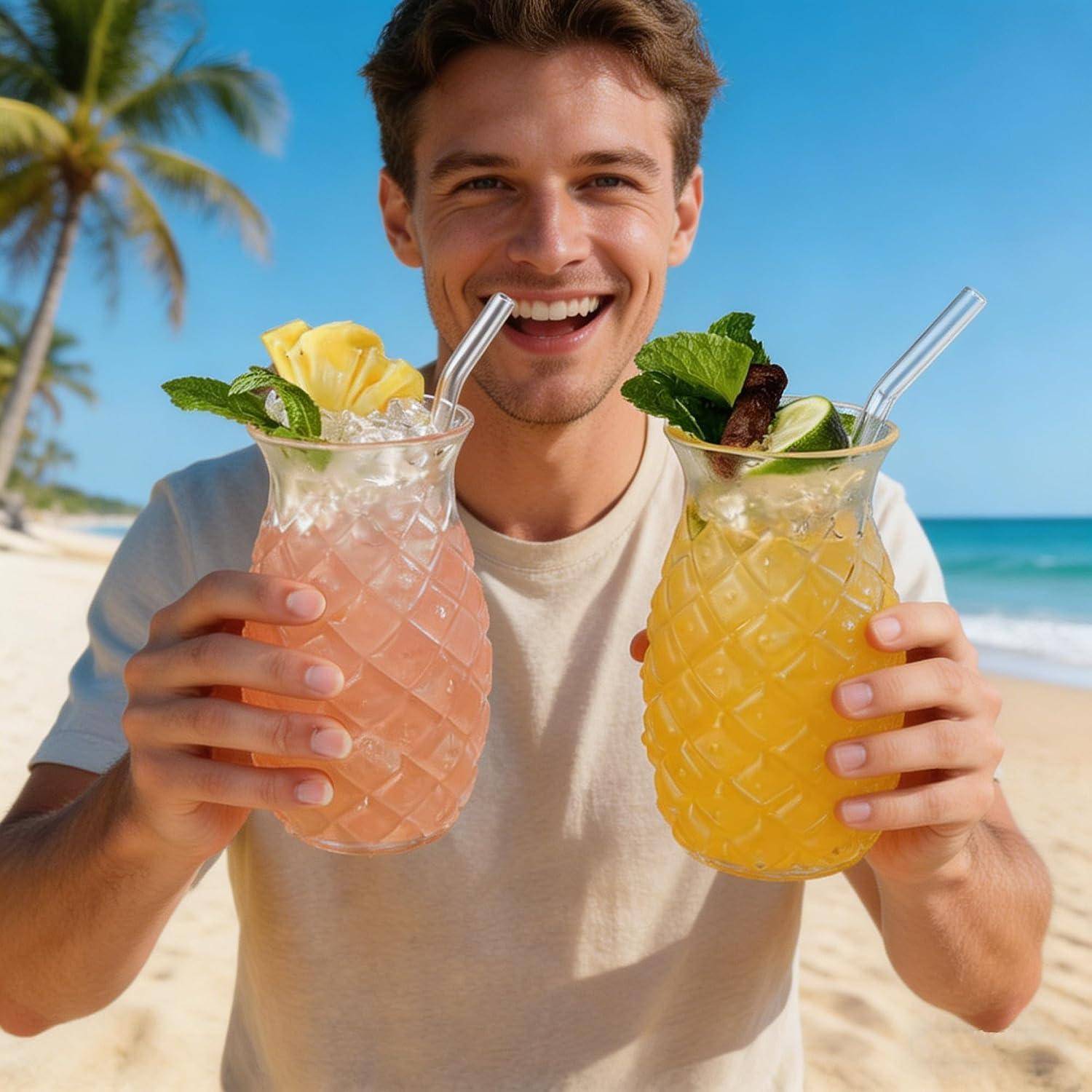 Ein lächelnder Mann an einem sonnigen Strand hält zwei tropische Cocktails in Glastassen mit Strohhalmen und Minzgarnitur hoch.