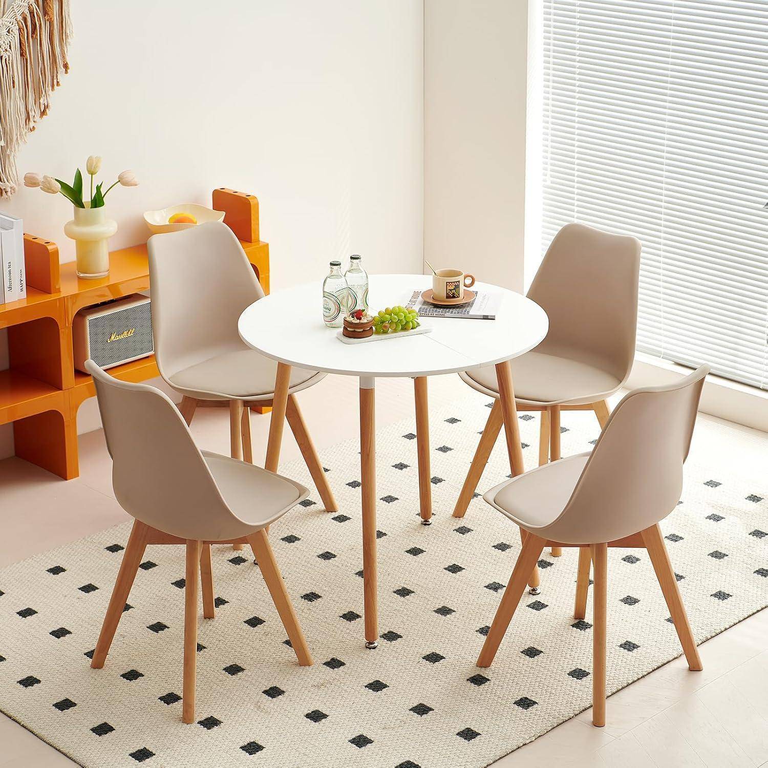 Kleiner moderner Essbereich mit einem runden weißen Tisch und vier beigefarbenen Stühlen auf einem gemusterten Teppich, neben einem hellen Fenster und orangefarbenen Regalen.