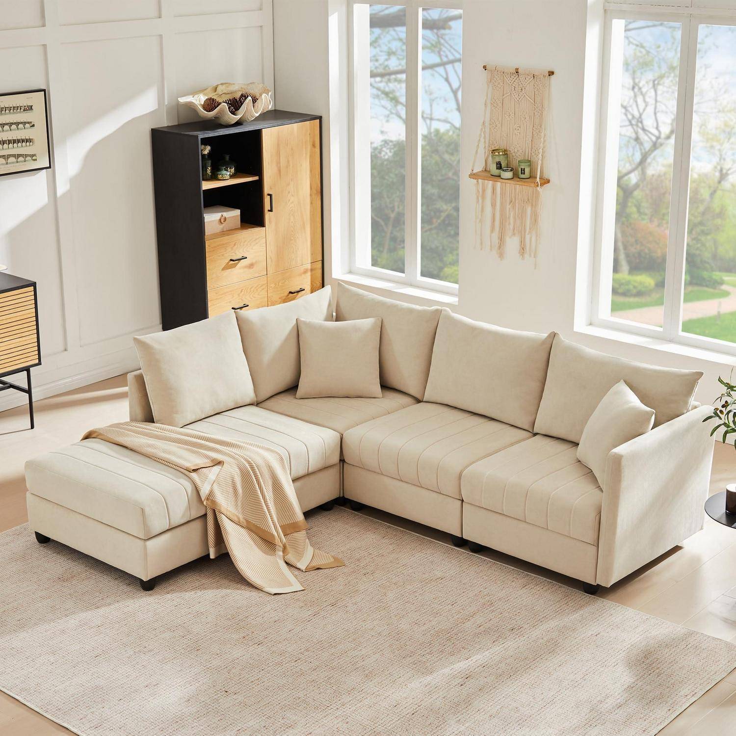 Beige L-förmiges Ecksofa mit Ottomane in einem hellen Wohnzimmer, gezeigt mit einer Wurfdecke, Kissen, Schrank und großen Fenstern.