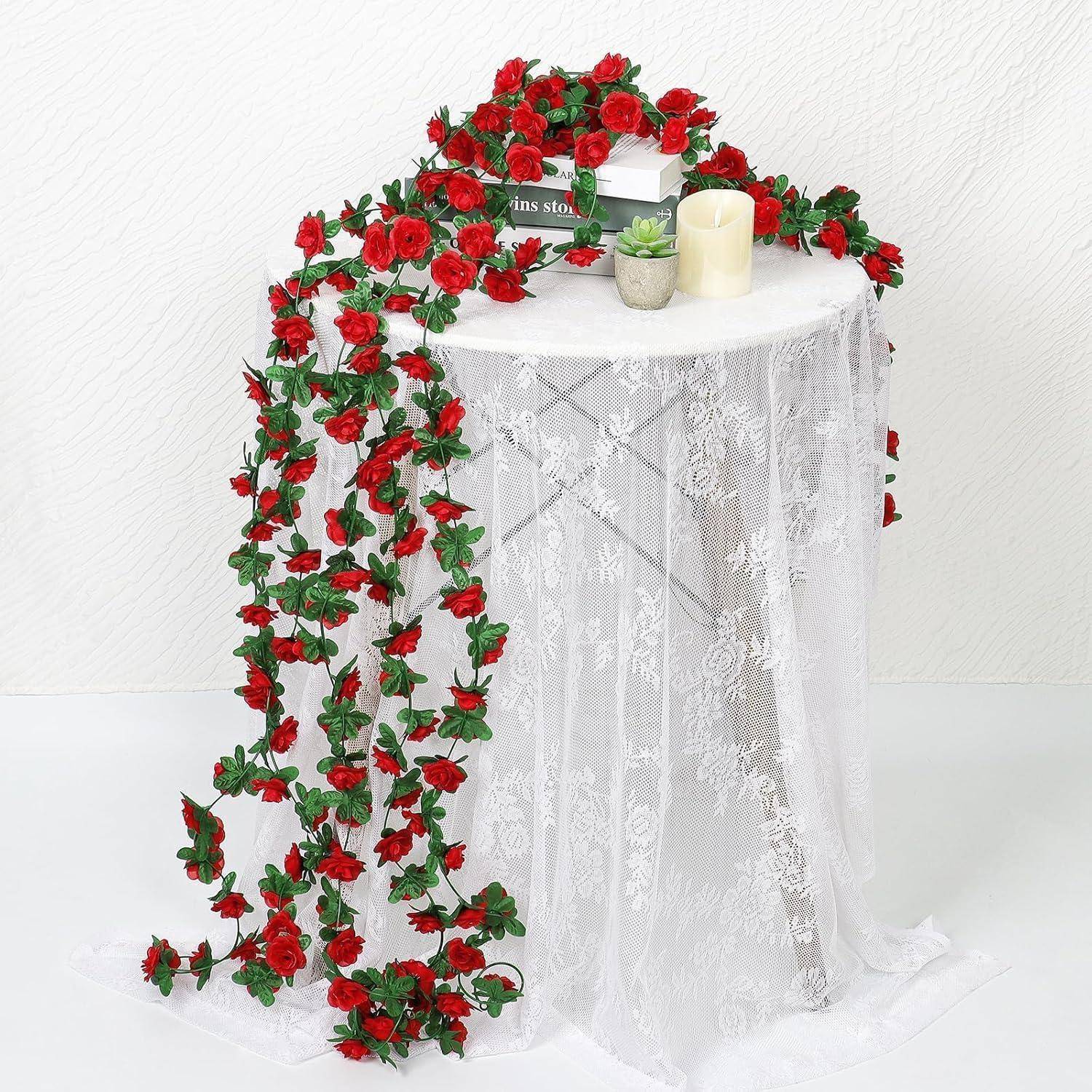 Weißer, mit Spitze bedeckter Säulentisch, dekoriert mit einer kaskadierenden Girlande aus roten Rosen, einer Kerze und einer kleinen Pflanze auf der Oberseite.