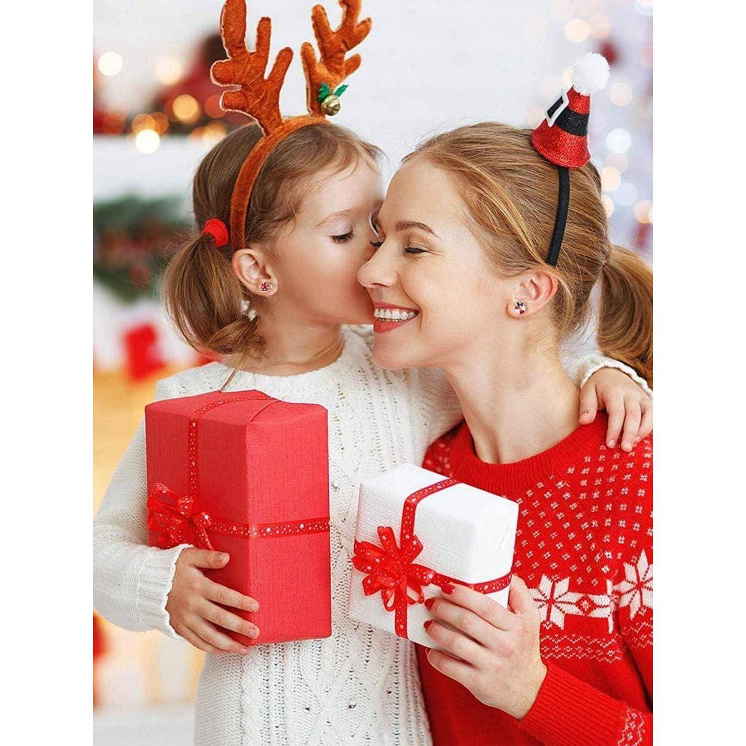 Junges Mädchen mit Rentiergeweihen umarmt und küsst eine lächelnde Frau in einem Weihnachtspullover; beide halten eingepackte Geschenke.