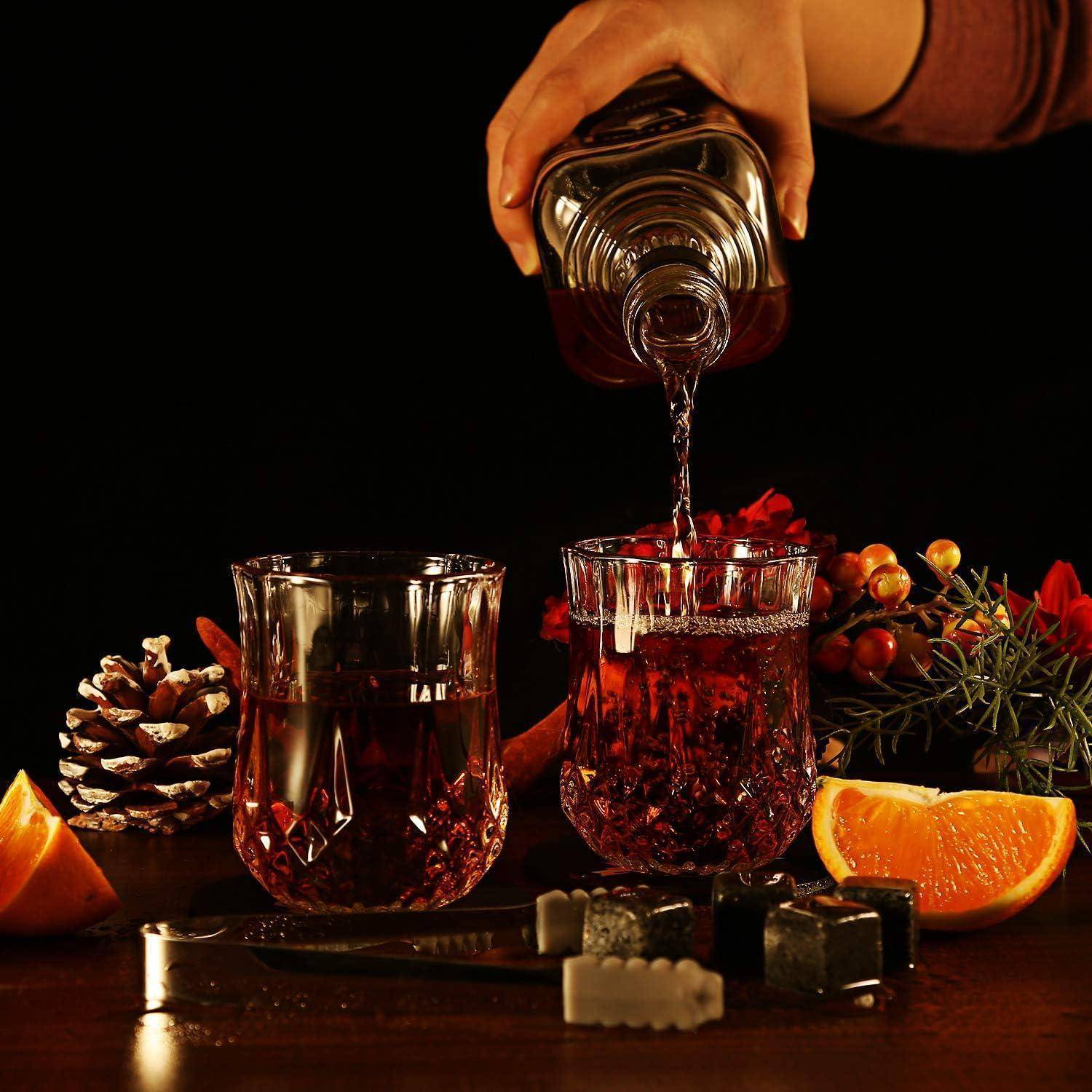 Eine Hand gießt ein rotes Getränk aus einer Flasche in ein Glas neben einem gefüllten Kristallglas, mit Orangenscheiben und Tannenzapfen auf einem dunklen Tisch.