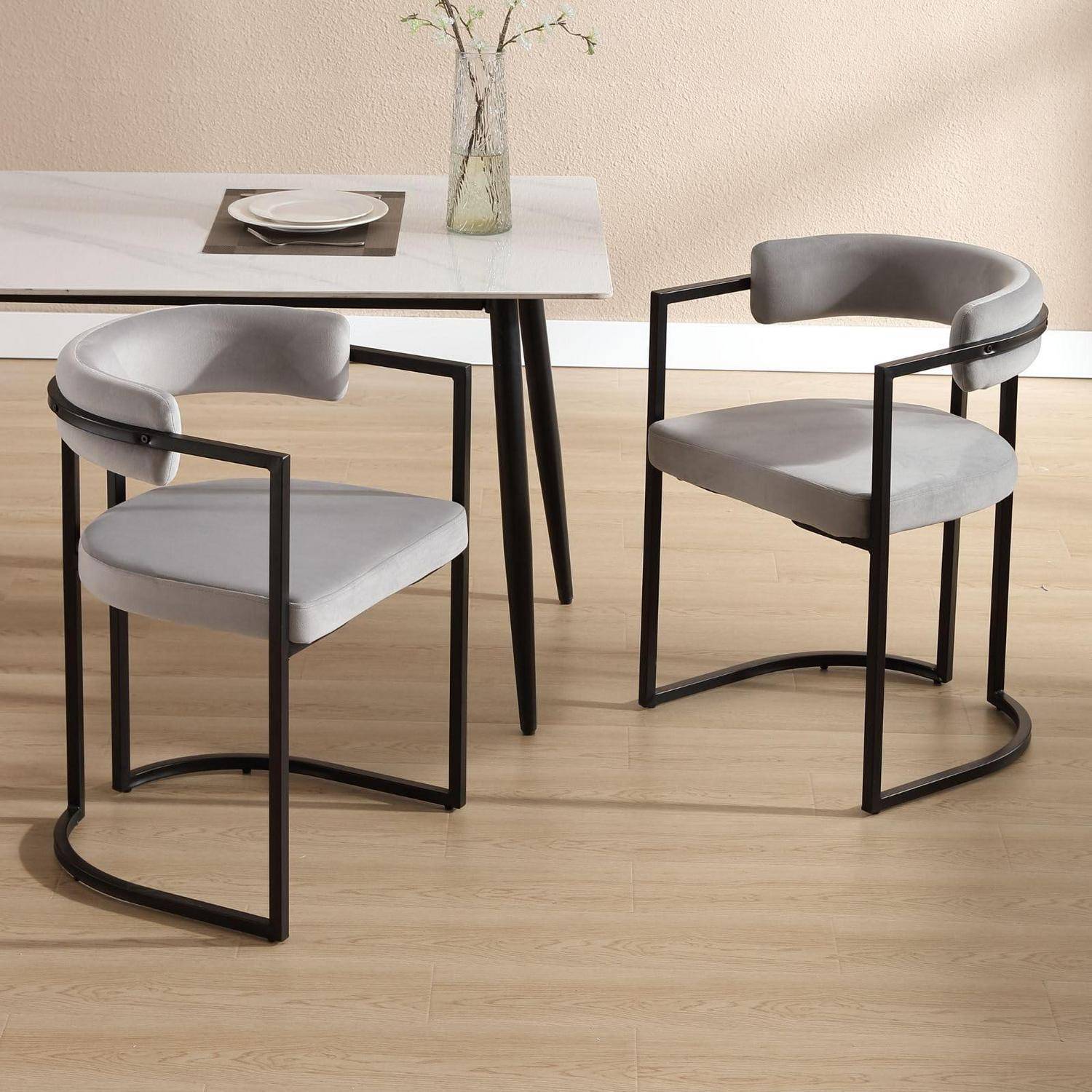 Zwei grau gepolsterte Esszimmerstühle mit schwarzen Metallrahmen stehen neben einem weißen Tisch, der mit Tellern und einer Vase in einem minimalistischen Raum eingedeckt ist.