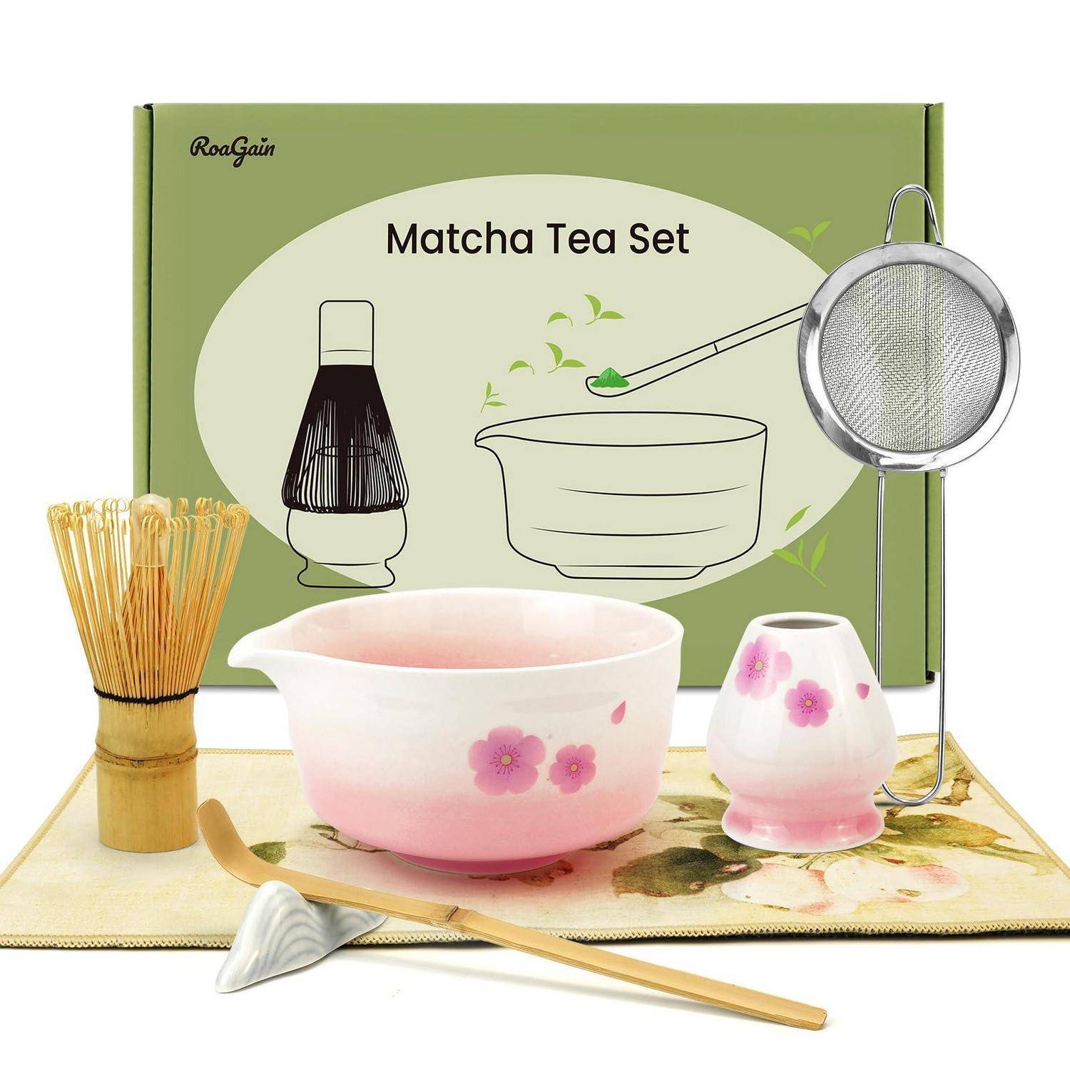 Matcha-Teeset mit rosafarbener Blütenschüssel, Besen, Löffel, Sieb und kleiner Tasse auf einer Matte, dargestellt in Produktverpackung.