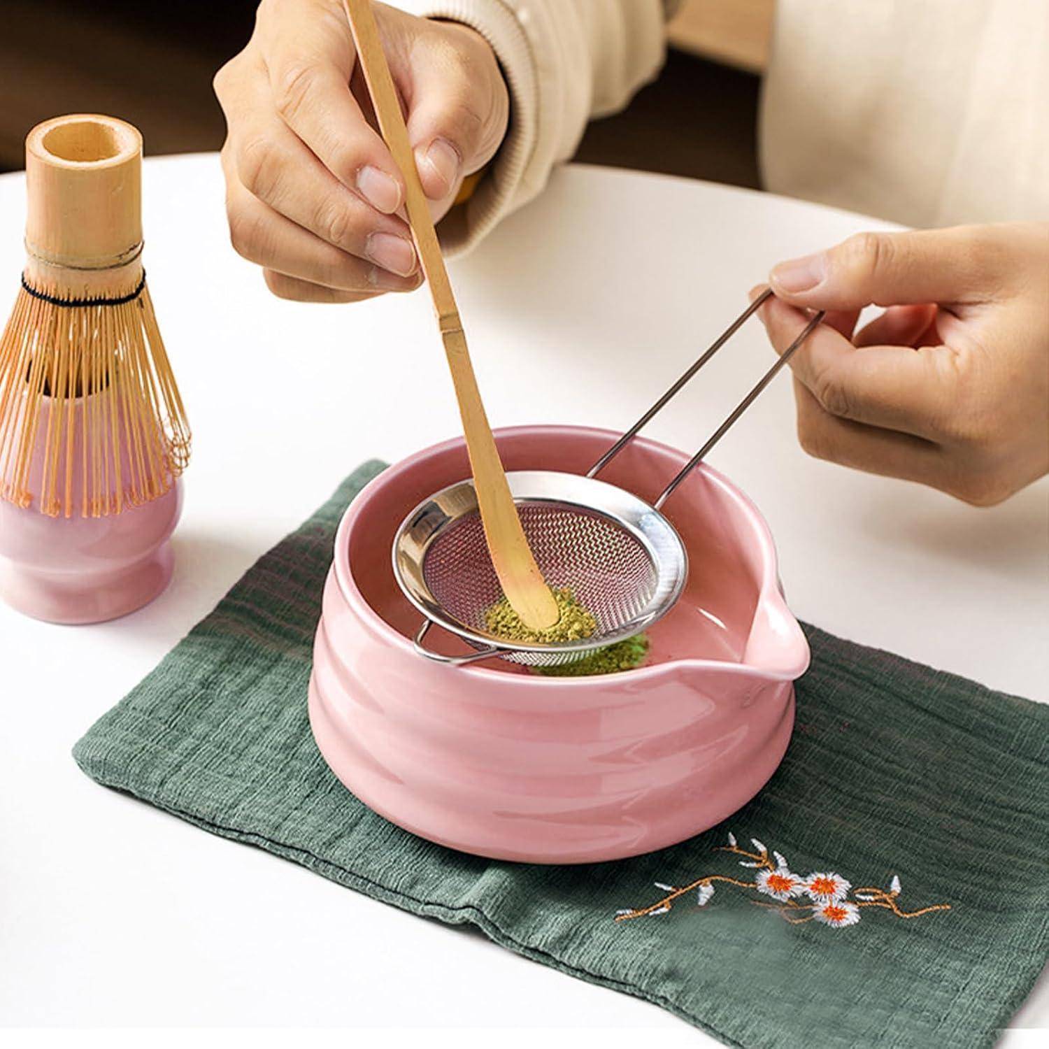 Hände bereiten Matcha-Tee zu, rühren das Pulver in einer rosa Schüssel mit einem Bambusbesen um und sieben es durch ein Maschensieb auf einer Tischunterlage.